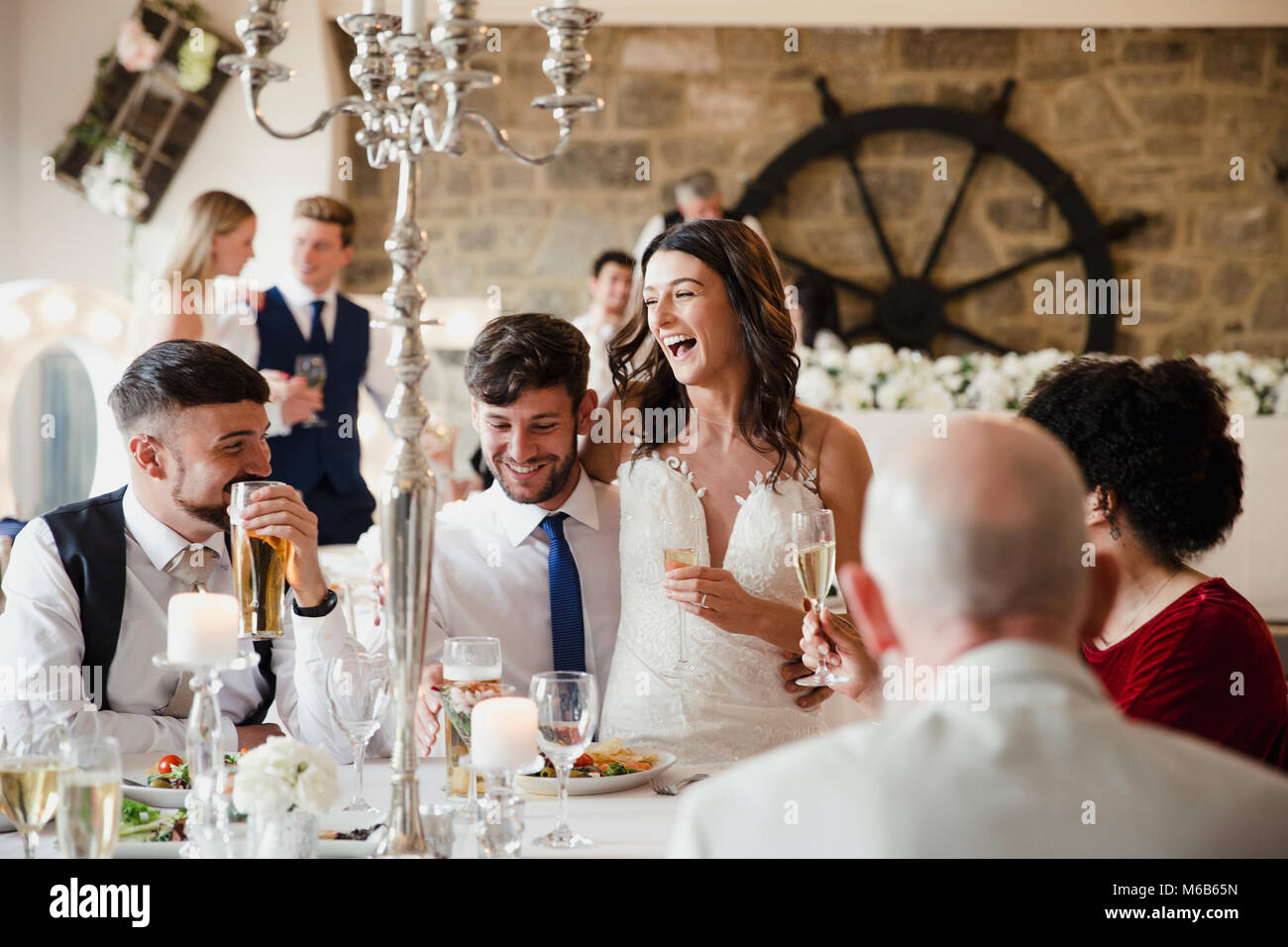 Heureux, couple de jeunes mariés sont socialiser avec les clients de leur repas de mariage. La mariée est assise sur les genoux de son mari et ils rient et drinki Banque D'Images
