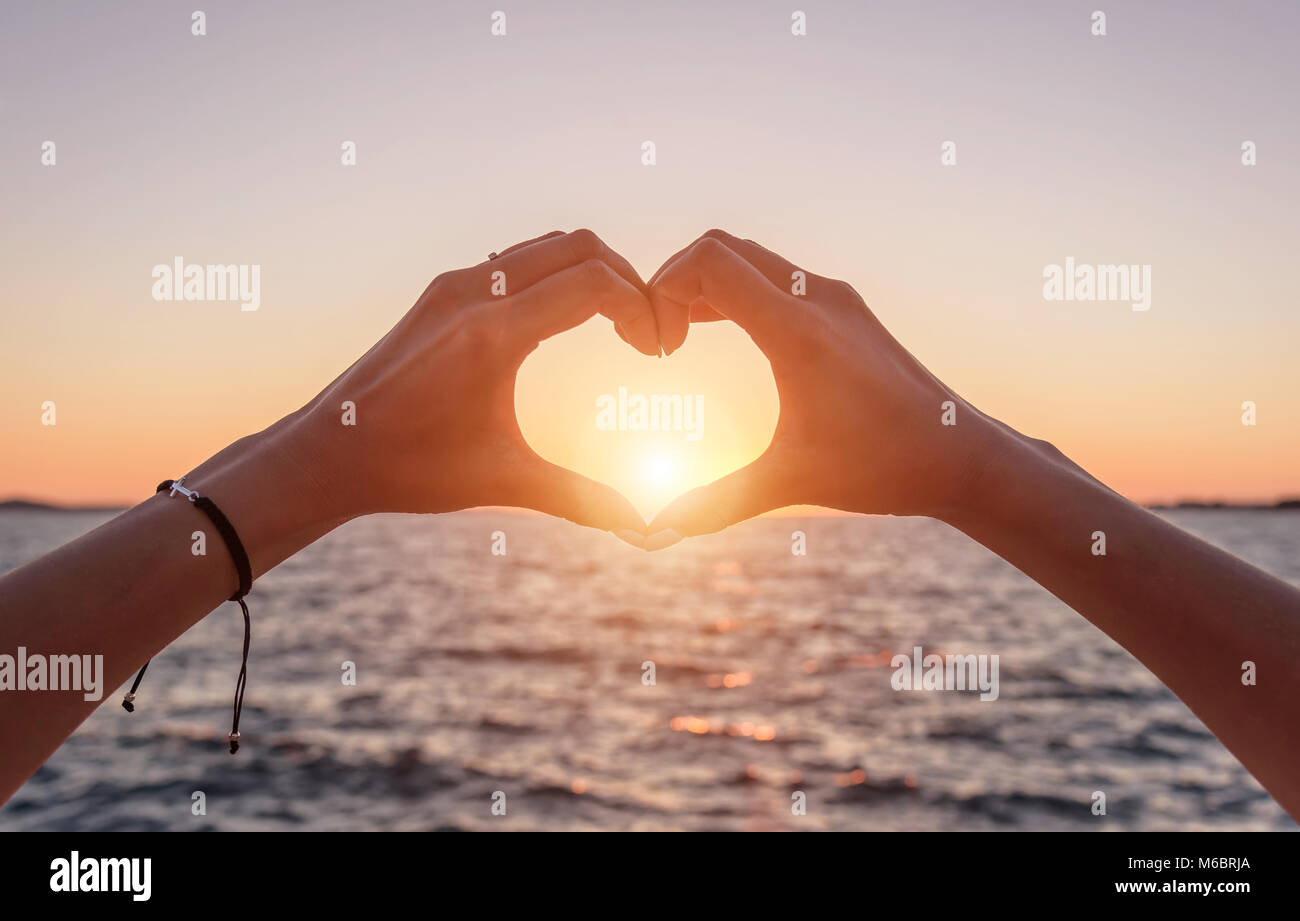 Mains en forme de cœur au coucher du soleil et contre le fond de la mer. Banque D'Images