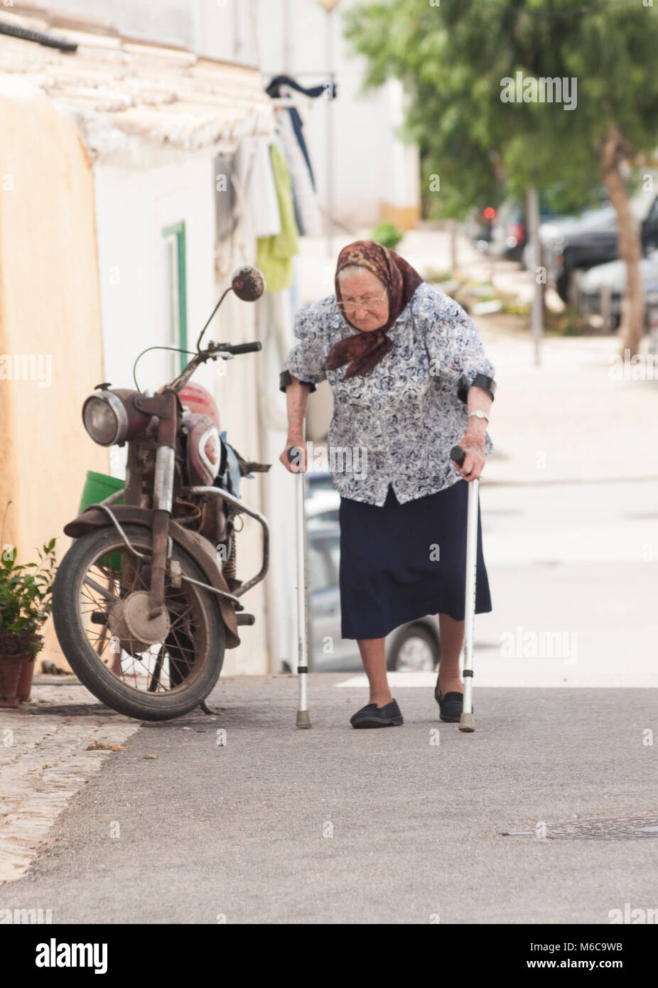 Femme plus âgée à béquilles aller marcher dans un petit village portugais. Une moto à l'arrière-plan. Banque D'Images
