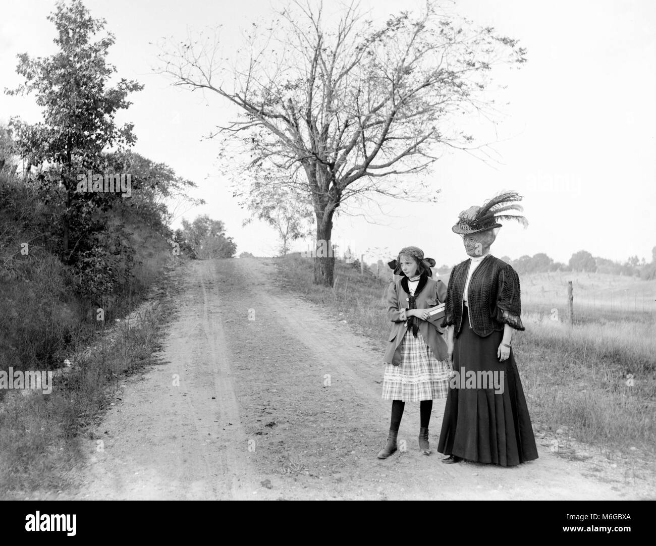 Un élégant à la grand-mère et sa petite-fille se prépare pour l'école et longue marche vers le bas une route de campagne, ca. 1900. Banque D'Images