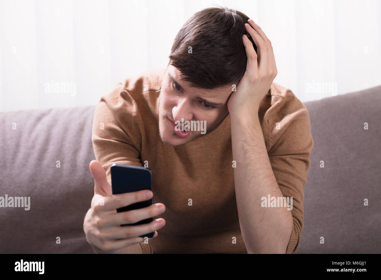 Choqué jeune homme assis sur un canapé et Looking At Smartphone à la maison Banque D'Images