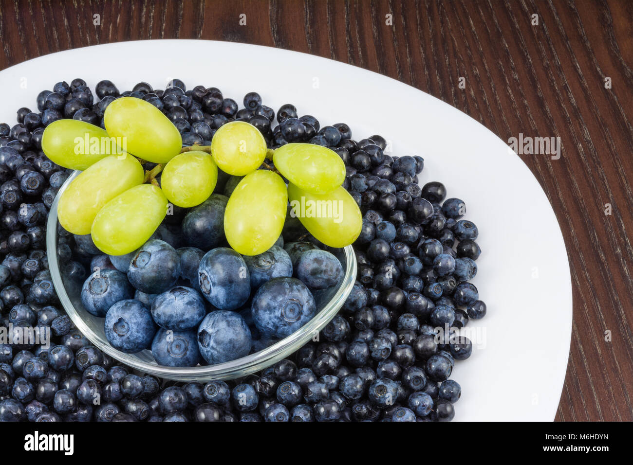 Les bleuets et grappe de raisins sur table. Vaccinium myrtillus. Close-up de plaque blanche avec des baies et bol en verre sur fond de bois. Banque D'Images