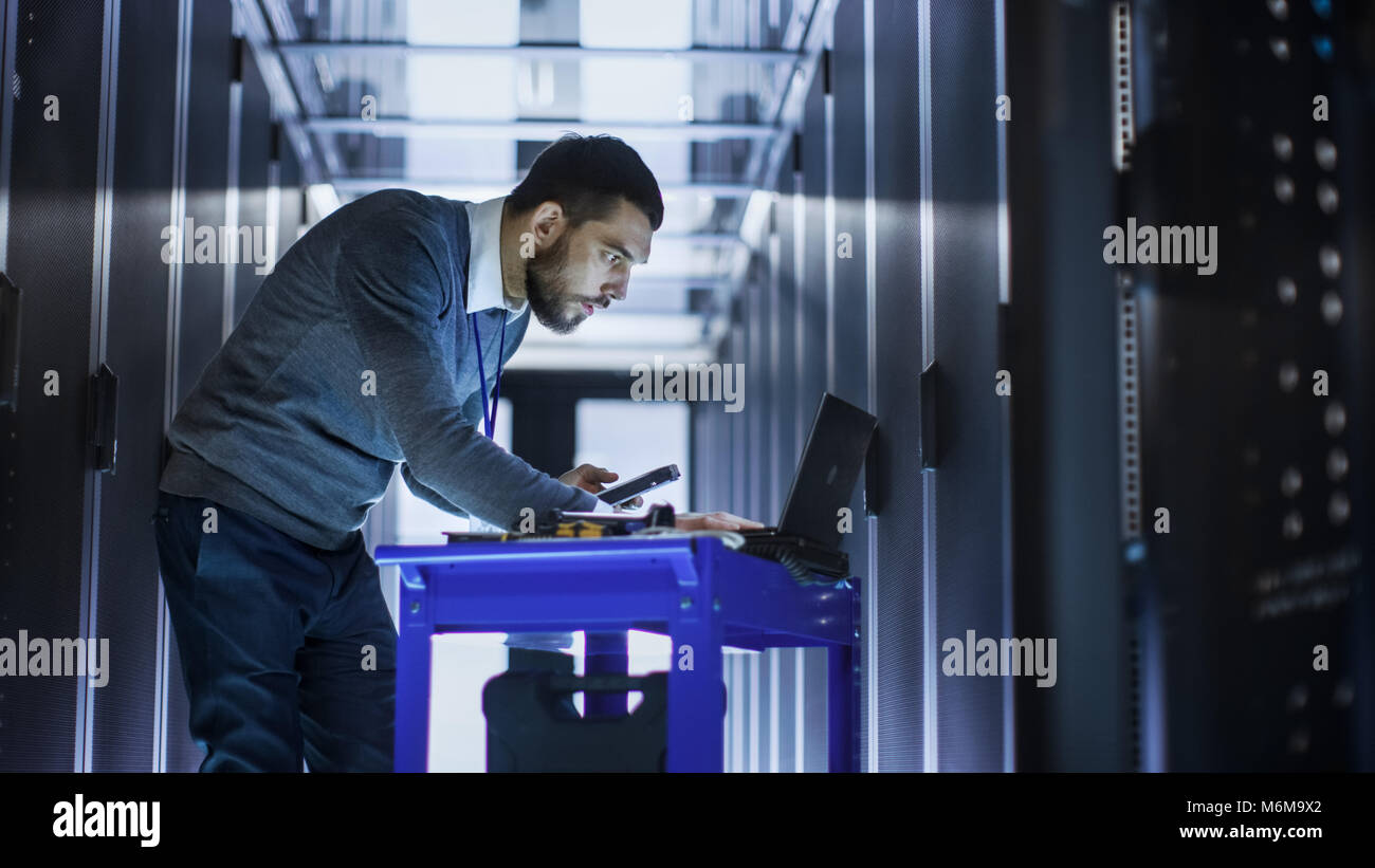 Ingénieur en Informatique avec l'outil Panier travaillant sur un ordinateur portable, il est titulaire d'un disque dur. Il se tient à un couloir d'un grand centre de données complet de serveurs en rack. Banque D'Images