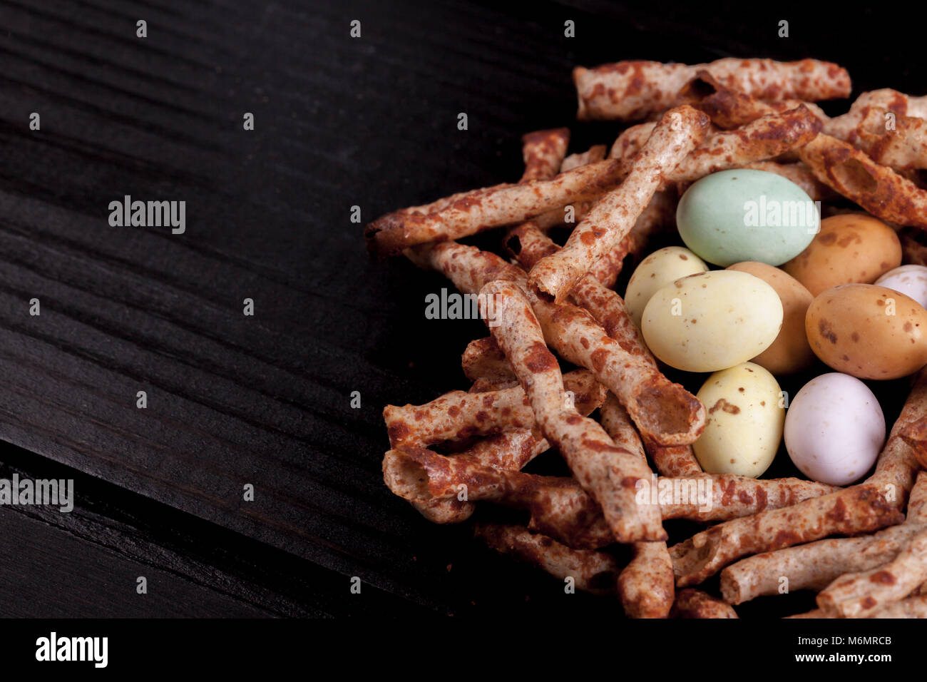 Nid de Pâques à base de chips comestibles qui ressemblent à des bâtons, et des bonbons au chocolat recouvert d'œufs. Les oiseaux nichent sur une table en bois noir avec copie espace Banque D'Images