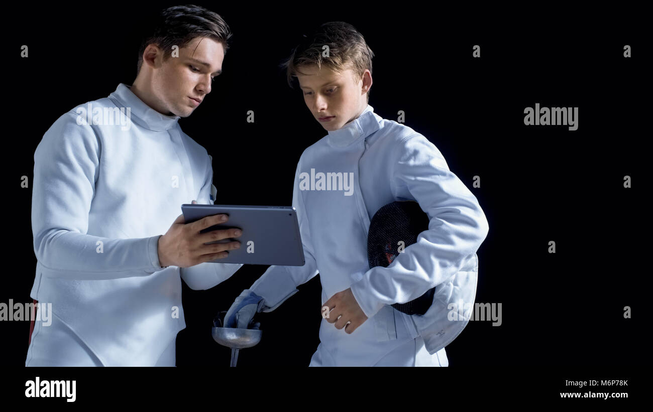 Deux jeunes tireurs d'utiliser l'ordinateur tablette pour en savoir plus sur la stratégie, l'attaque et la défense dans l'escrime. Shot isolé sur fond noir. Banque D'Images