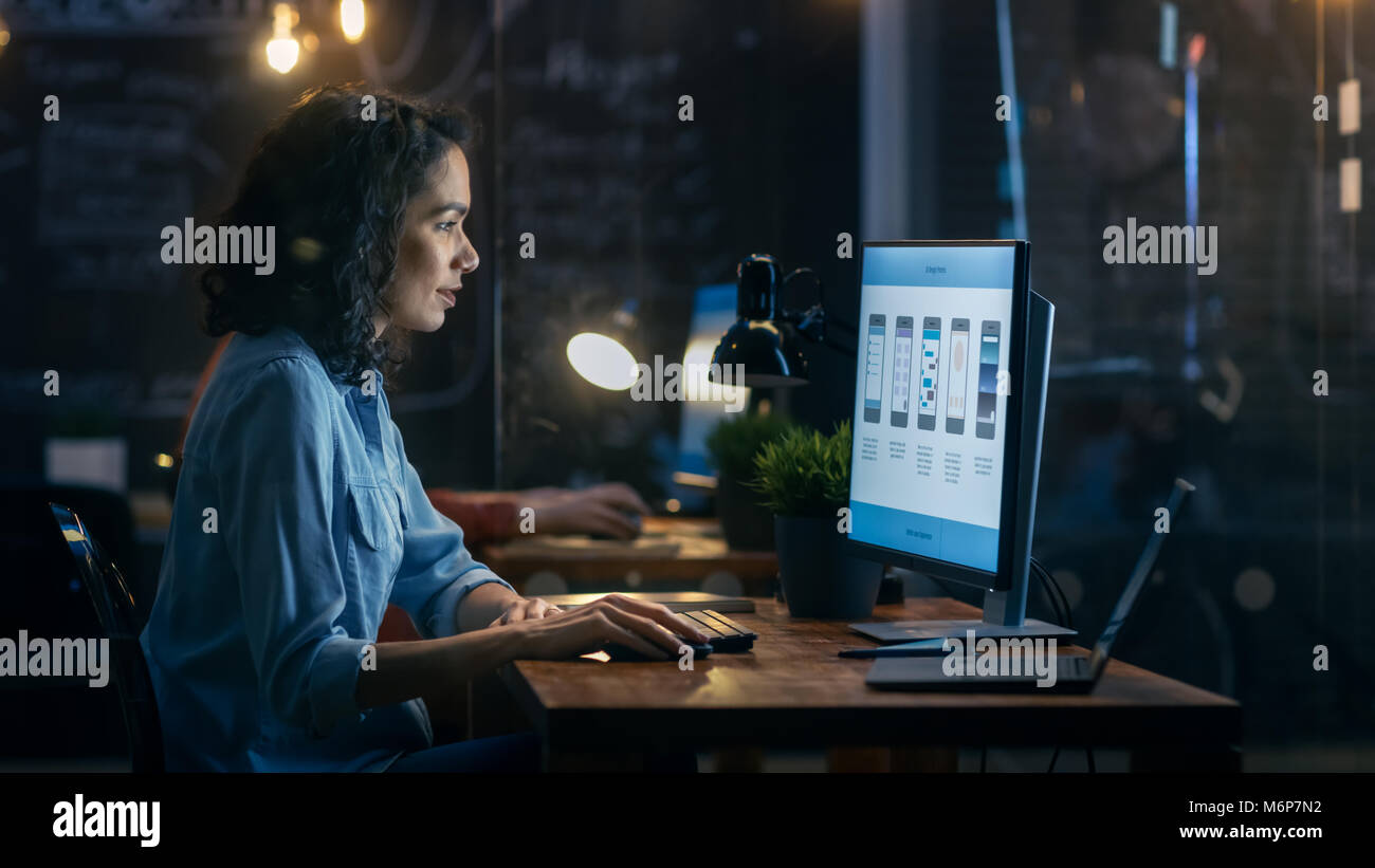Belle femme Coder travaille à son bureau sur son ordinateur. Peropnal Son collègue masculin est assis à côté d'elle. Office du soir a Creative l'éclairage. Banque D'Images
