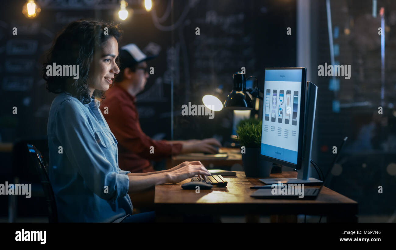 Belle femme Coder travaille à son bureau sur son ordinateur. Peropnal Son collègue masculin est assis à côté d'elle. Office du soir a Creative l'éclairage. Banque D'Images
