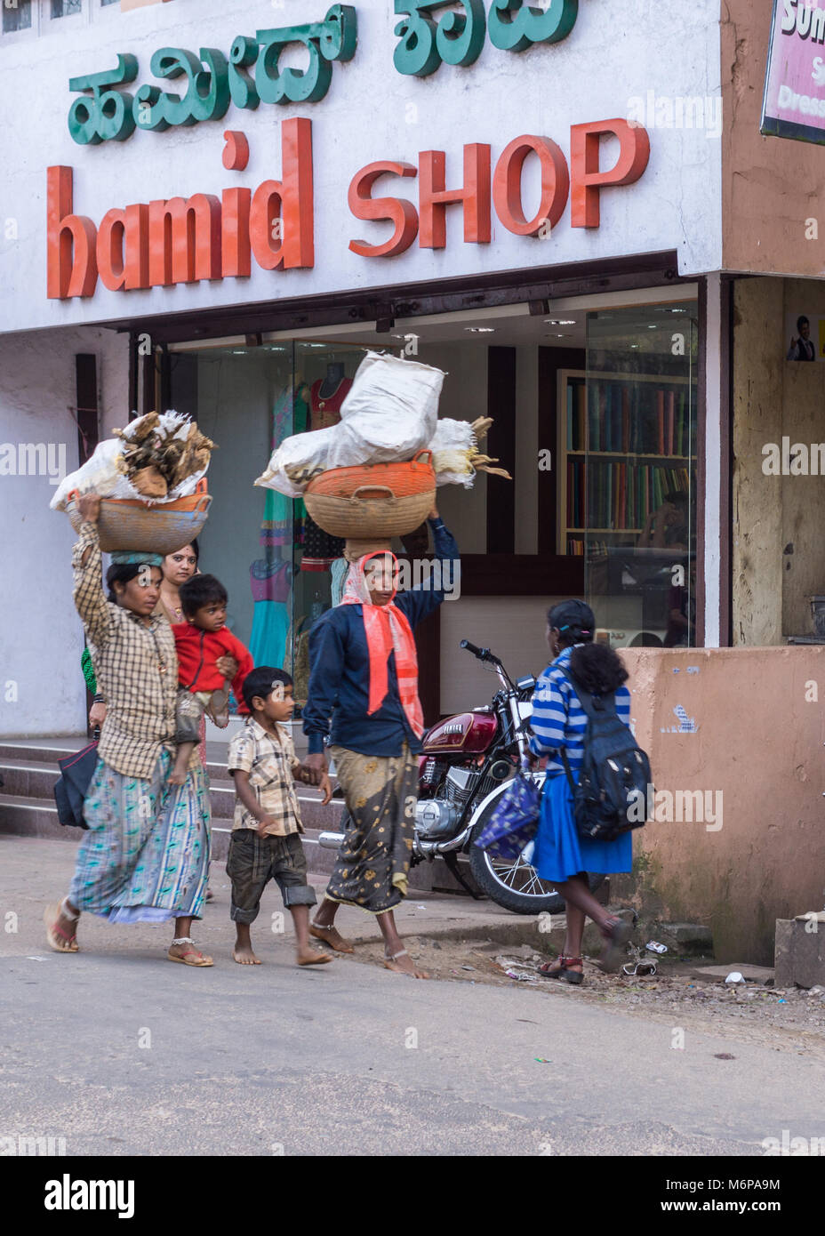 Madikeri, Inde - le 31 octobre 2013:Deux femmes avec enfants transporter le bois dans de grands paniers sur leurs têtes en face de boutique dans une scène de rue. Banque D'Images