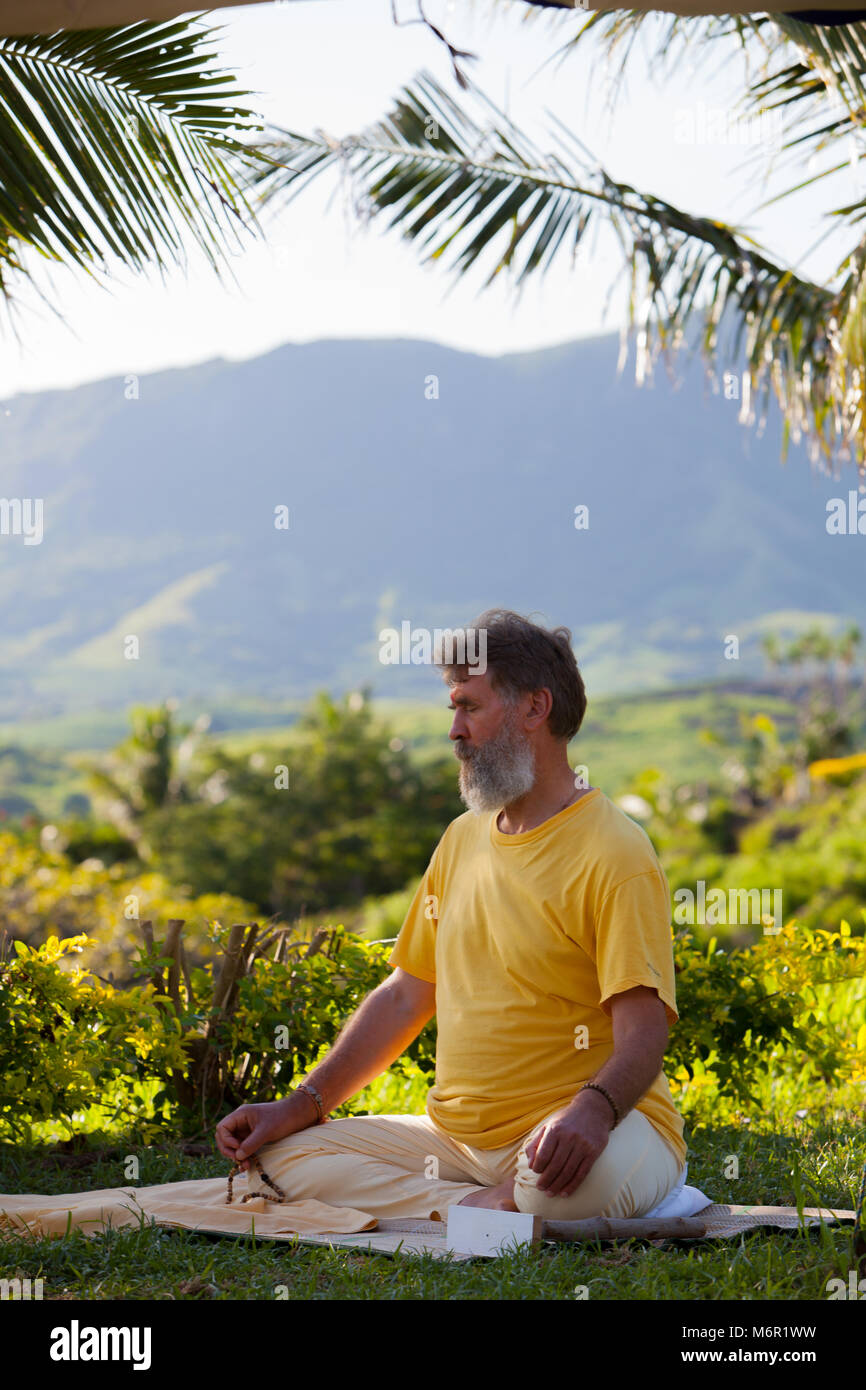 Un homme à barbe, assis sur un tapis de yoga et pratiquer la méditation avec perles mala dans la nature. Participation à battre en retraite dans les îles Fidji. Banque D'Images