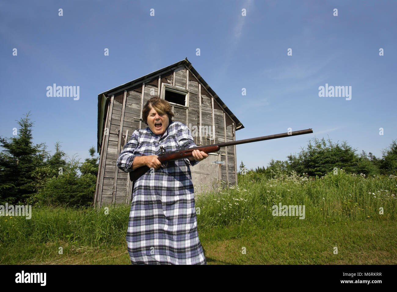 Photo d'une femme dans la soixantaine se balançant autour d'un fusil plutôt grand en face d'une ancienne grange. Banque D'Images
