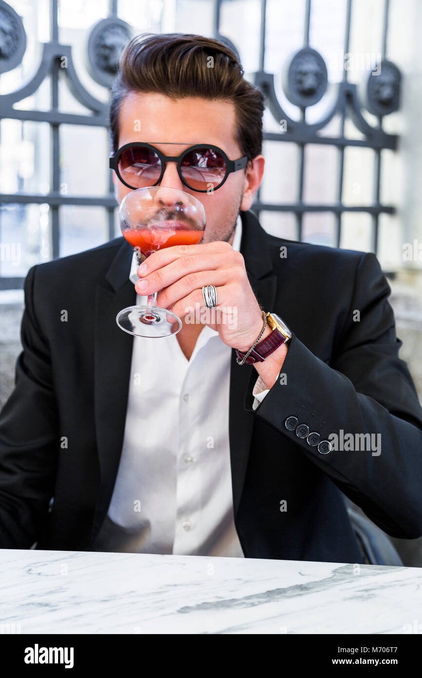 Beau jeune homme en costume noir avec chemise blanche, bracelets et  accessoires sur les mains dans des lunettes de soleil et cocktail rose  rouge potable assis dehors sur un Photo Stock -
