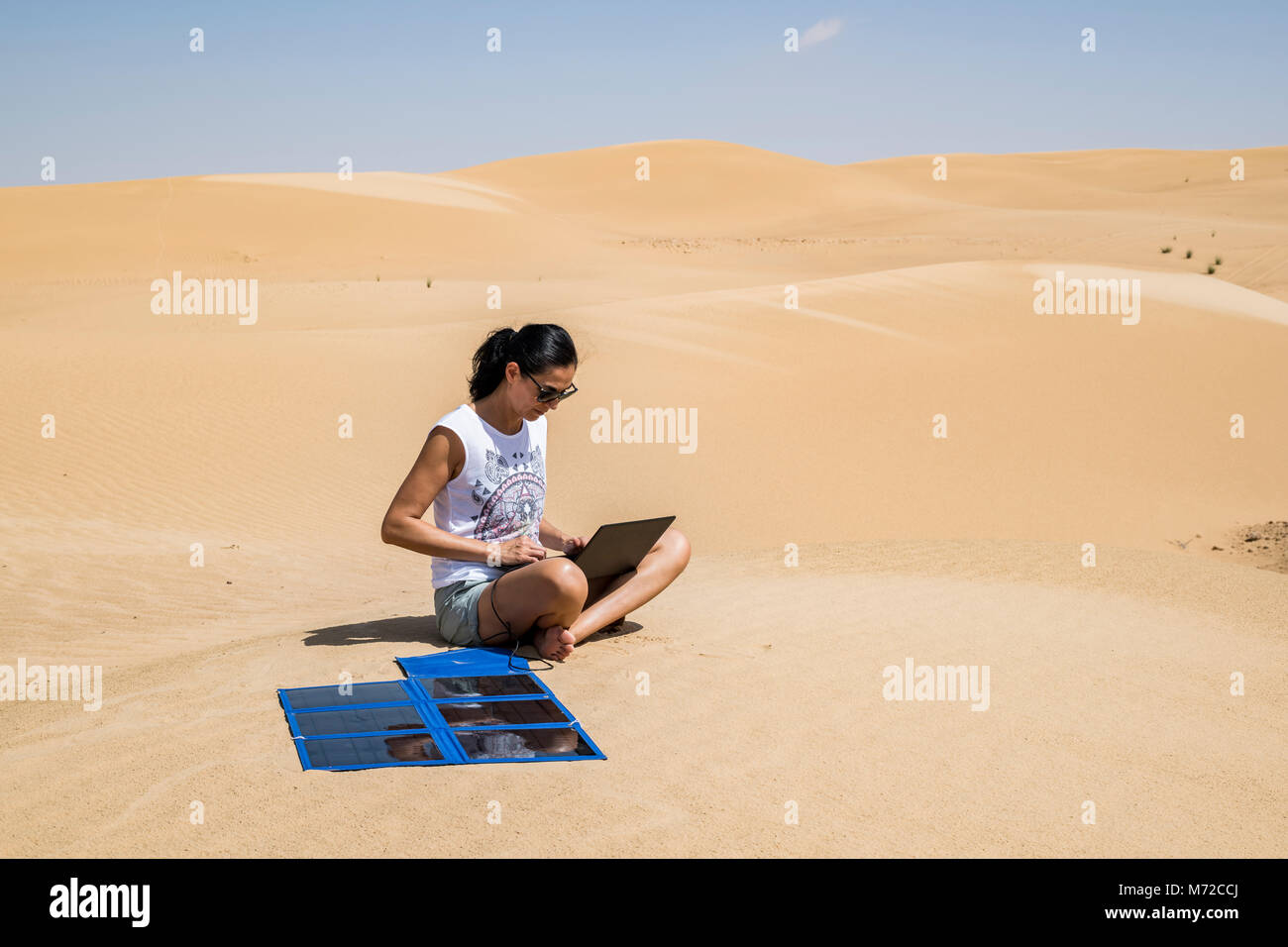 Femme adulte de l'aventurier au milieu des dunes en utilisant son ordinateur pendant la charge avec portable solar panneaux flexibles Banque D'Images