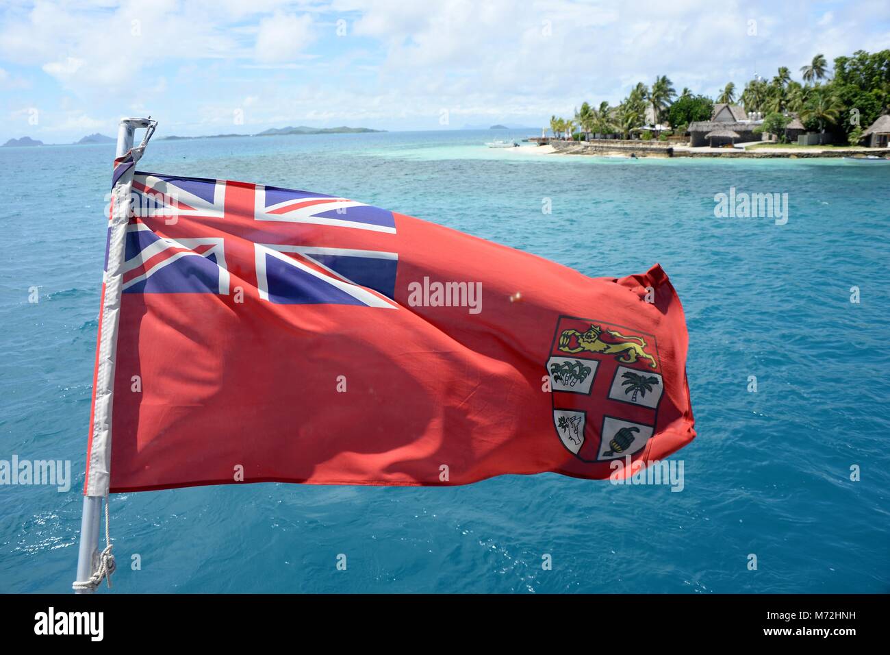 Le drapeau fidjien coups dans le vent à l'arrière d'un bateau la visite d'une belle île tropicale au large des Fidji. C'est le commerçant ensign avec un fond rouge. Il y a un gouvernement ensign avec un fond bleu qui représente la mer bleue de l'océan Pacifique. Il montre l'Union Jack comme un signe de l'île, des liens étroits avec la Grande-Bretagne. Banque D'Images