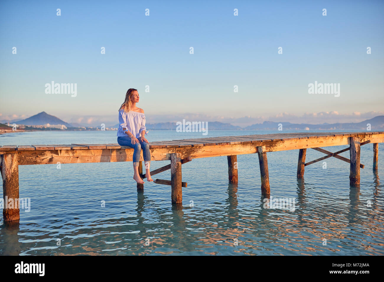 Nu femme assise sur une jetée en bois rustique ou promenade côtière au coucher du soleil avec ses jambes se balançant au-dessus de la mer vers le soleil couchant Banque D'Images