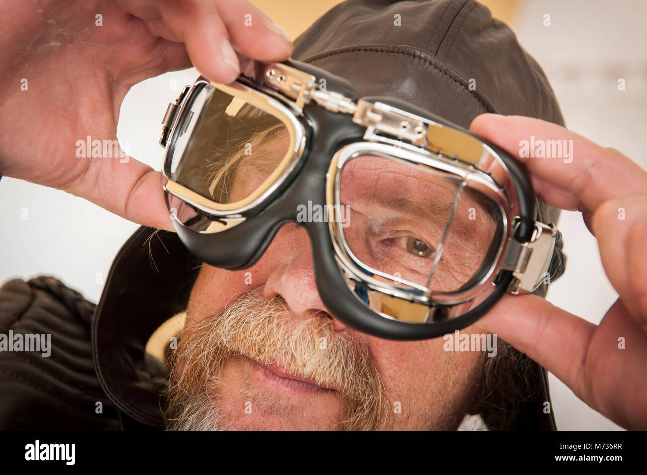 Close-up of a senior man avec barbe, chapeau de cuir tient avec les deux mains un lunettes moto en face de son visage et des clins d'dans l'appareil photo Banque D'Images