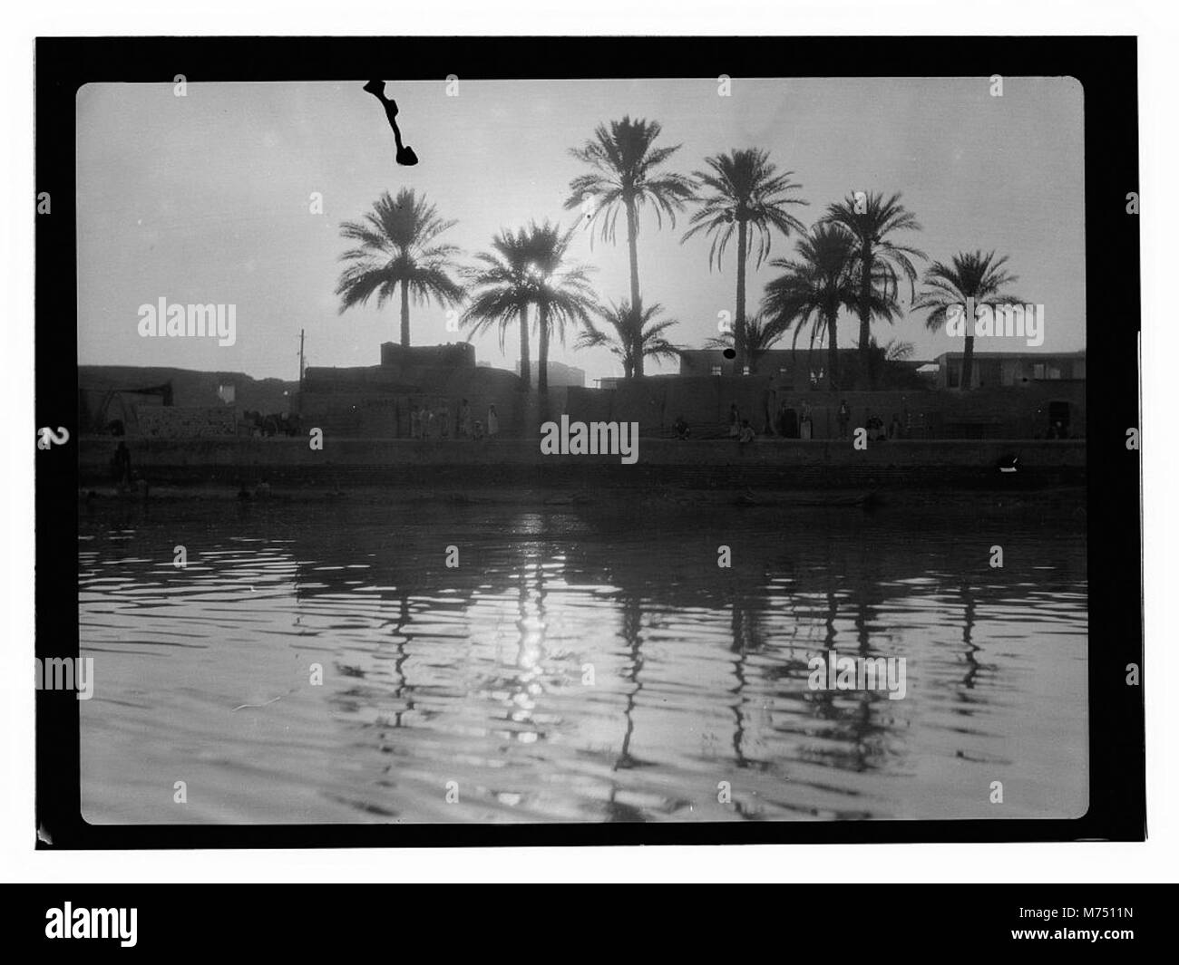 Une photographie de personnes utilisant des bateaux sur une rivière en Irak, avec une ville visible sur la rive opposée. Cette scène capture la vie quotidienne le long de la rivière, mettant en évidence les transports locaux et la vie communautaire dans la région. Banque D'Images