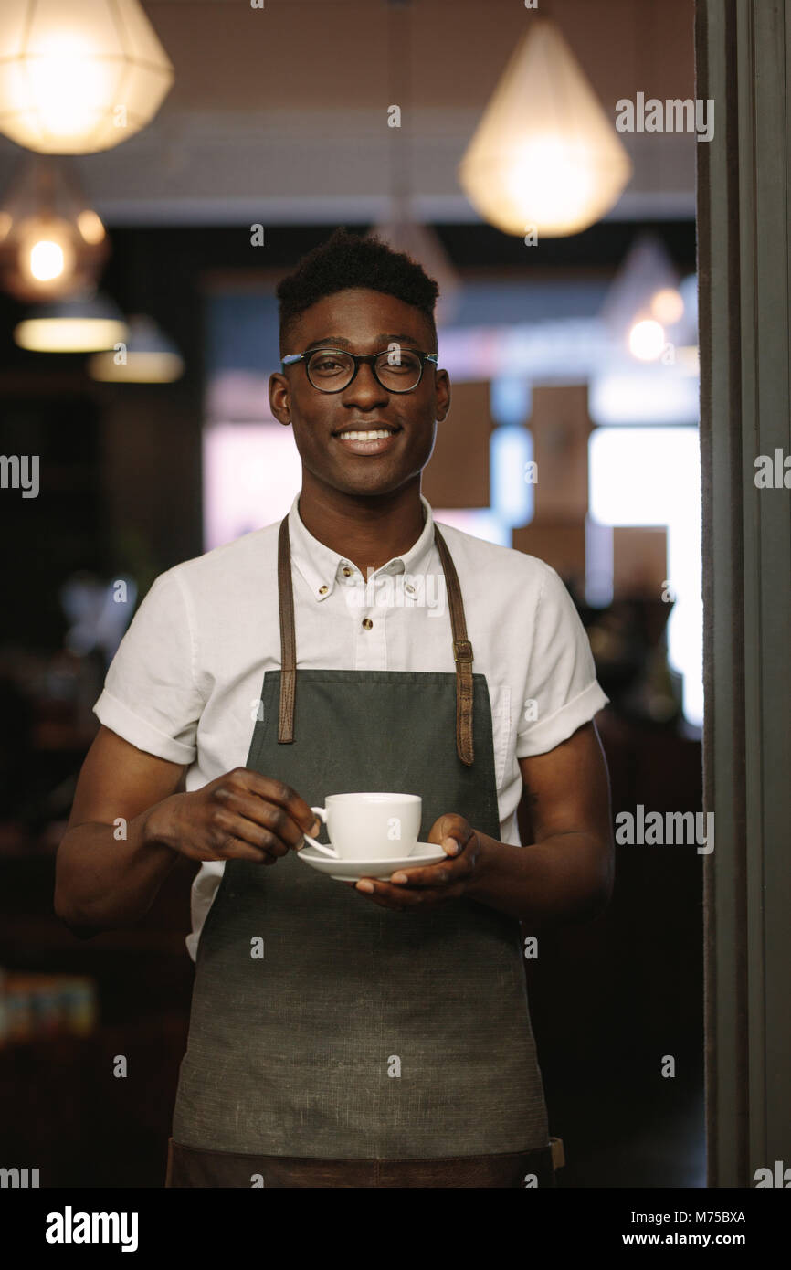 Gros plan du barista debout à l'entrée de café tenant une tasse de café. Heureux propriétaire d'une boutique de café posant avec une tasse de café dans la main. Banque D'Images