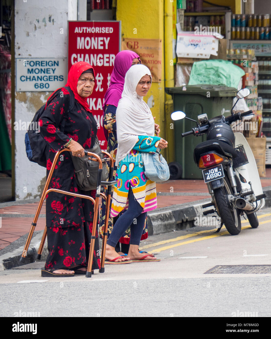 Deux jeunes femmes, et une femme âgée à l'aide d'un dispositif d'aide à la marche, traversant la rue à Singapour. Banque D'Images