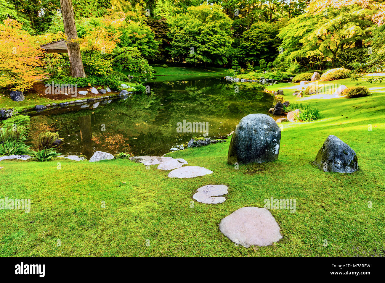 Une pelouse avec de l'herbe verte coupée près d'une rivière dans un parc avec des arbres et arbustes, pierres et un gazebo en bois sur une journée ensoleillée Banque D'Images