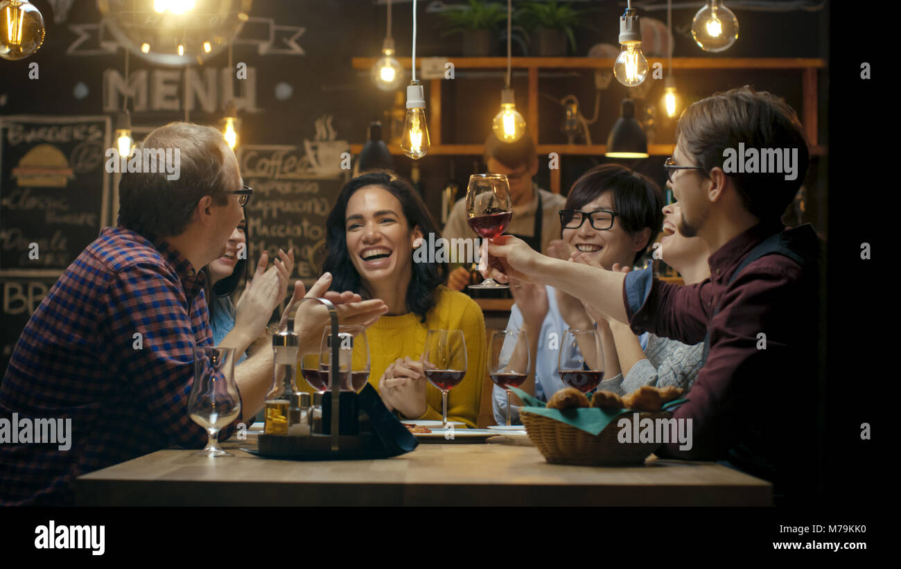 Groupe diversifié d'amis célébrer avec un toast et soulever des verres à vin en fête. Beaux jeunes gens s'amuser dans l''élégant bar/ restaurant. Banque D'Images