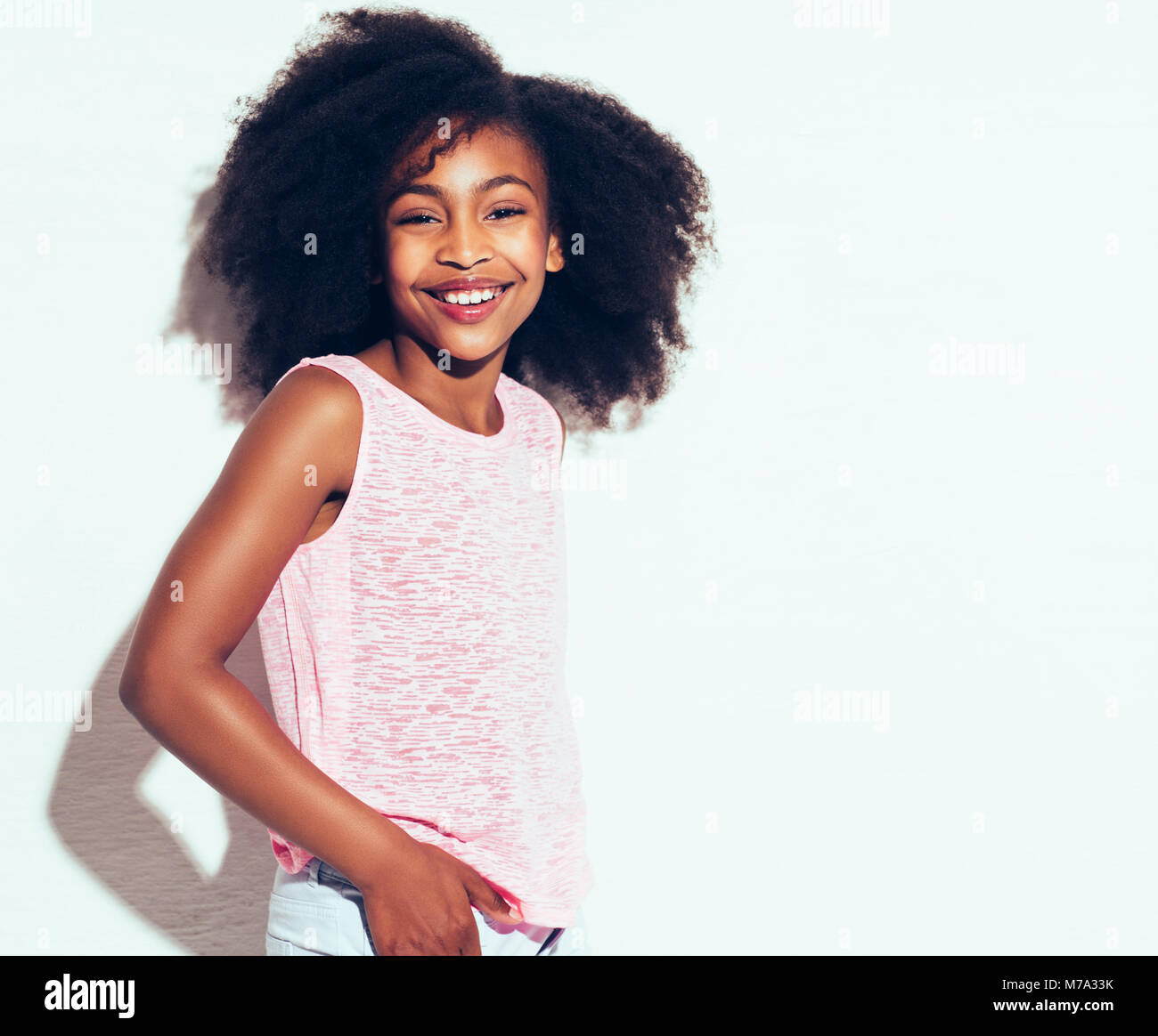 Cheerful young African girl avec de longs cheveux bouclés avec confiance tout en souriant tout seul sur un fond blanc. Banque D'Images