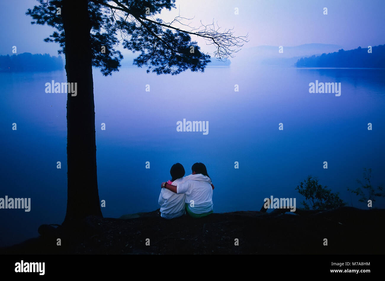 Deux filles préadolescentes siéger ensemble hugging regardant le coucher du soleil sur un lac dans le Vermont, USA. Banque D'Images