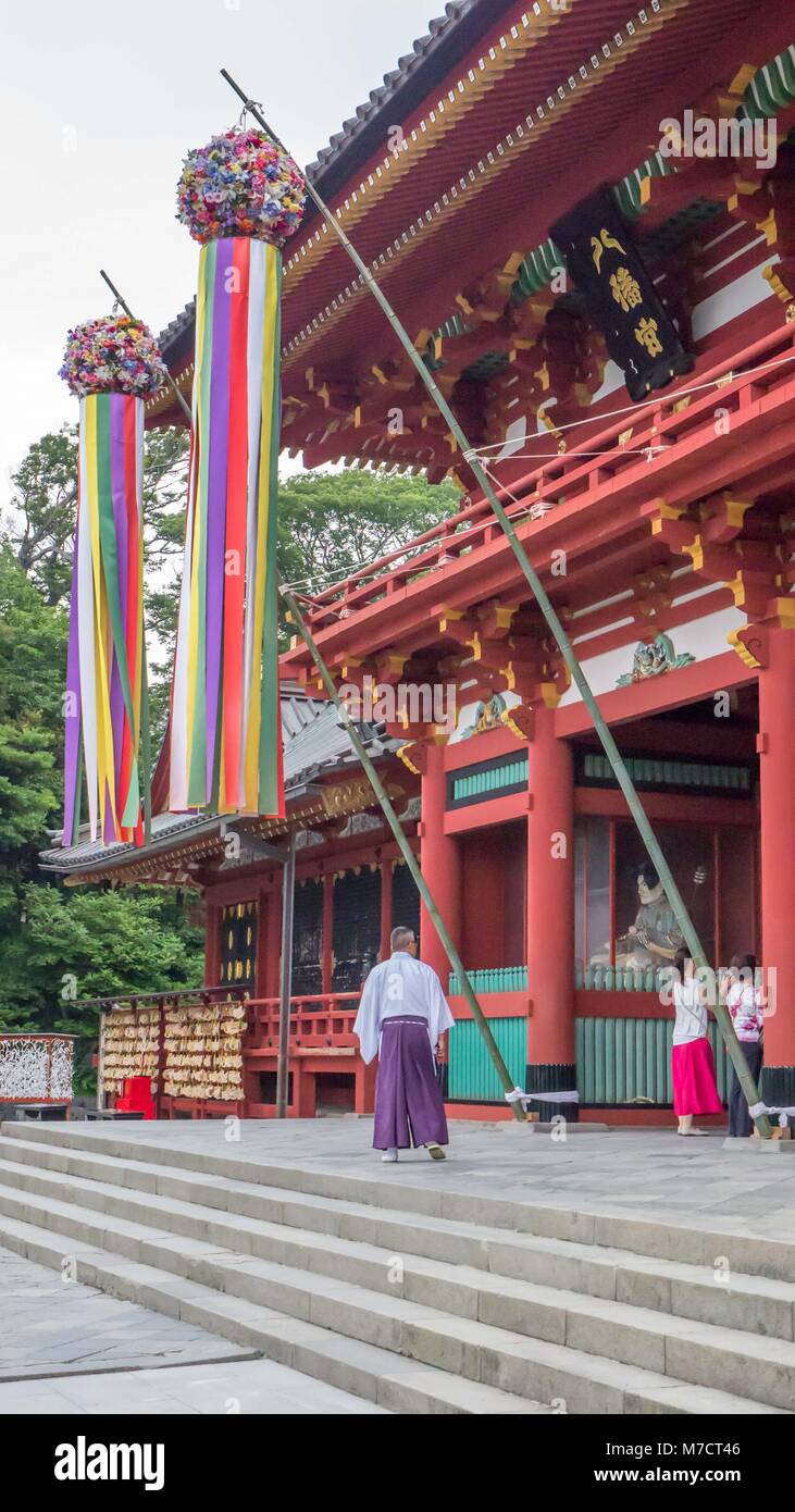 Prêtre Shinto habillé en blanc haut et pantalon violet à l'entrée du magnifique sanctuaire Tsurugaoka rouge dans Hachimangū avec de grandes décorations colorées. Banque D'Images