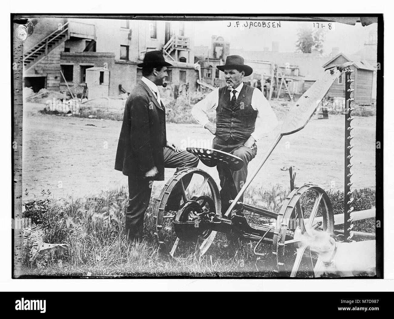 J.F. Jacobson se tient aux côtés d'un agriculteur et de matériel agricole dans une cour rurale, capturant une scène de la vie agricole au début du XXe siècle. Banque D'Images