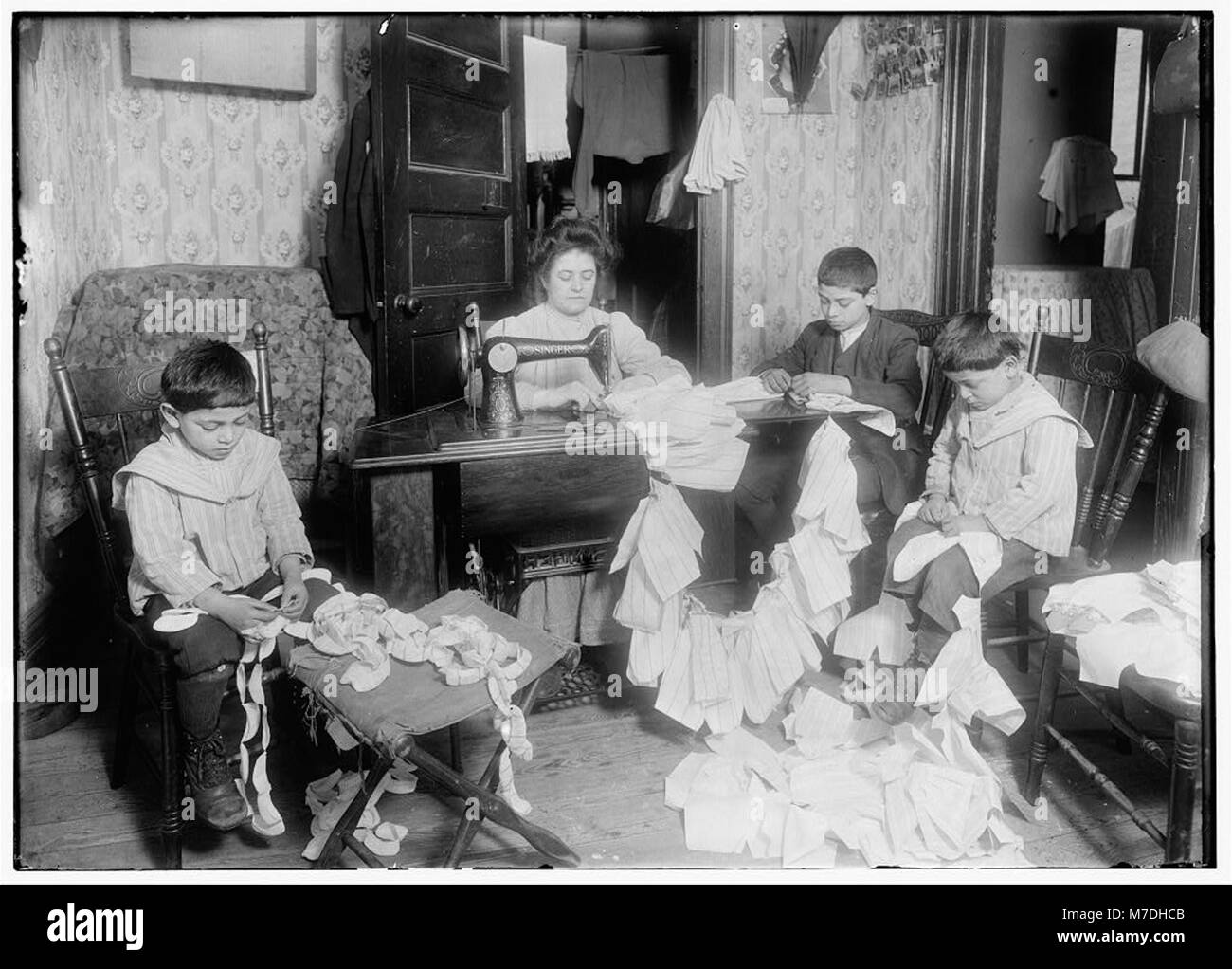 Cette photographie montre une famille dans une pièce exiguë au 4e étage du 59 Thompson Street, New York. La mère et son fils, âgés d'environ 12 ans, confectionnent des robes pour Campbell Kid Dolls, reflétant les conditions de vie difficiles du début du XXe siècle à New York. Banque D'Images