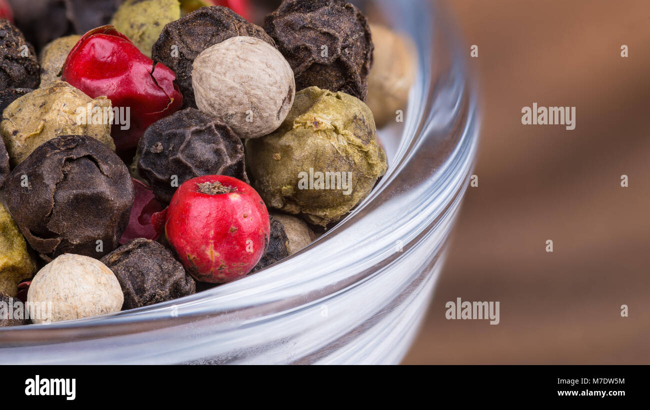 Close-up de différents grains de poivre dans un bol. Piper nigrum. Mélange coloré décoratif de poivre en grains secs dans le verre avec fond bois brouillée. Banque D'Images