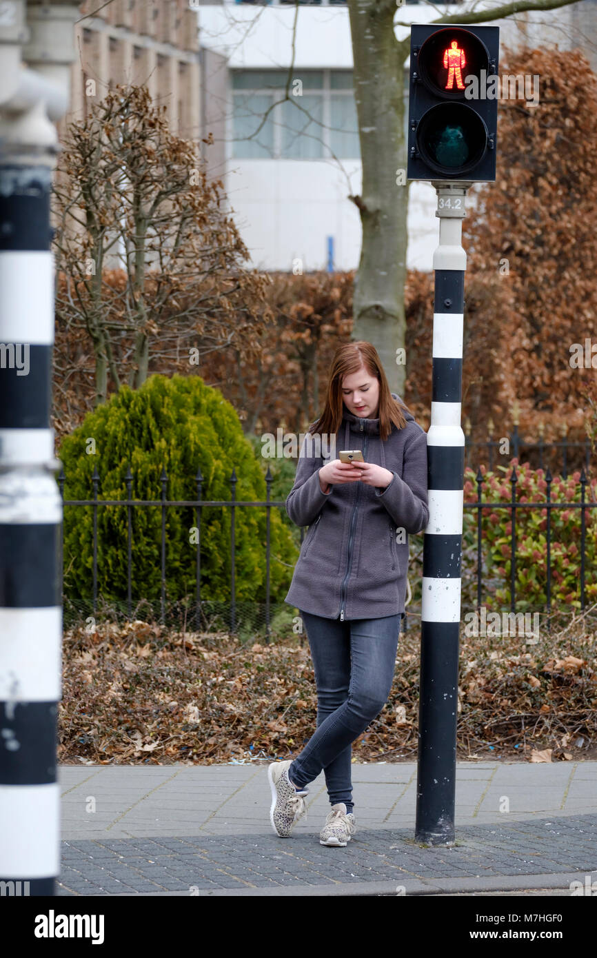 Jeune femme attend à un feu rouge qui est sur le rouge, mais ne fait pas attention à la circulation parce qu'elle regarde son téléphone mobile. Banque D'Images