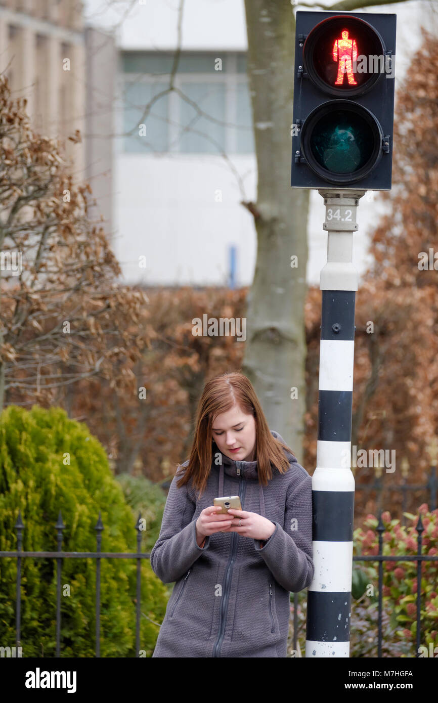 Jeune femme attend à un feu rouge qui est sur le rouge, mais ne fait pas attention à la circulation parce qu'elle regarde son téléphone mobile. Banque D'Images