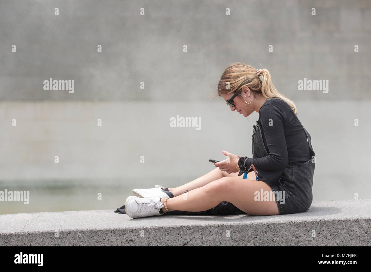Jeune femme blonde avec queue de cheval, assis sur le mur avec un téléphone mobile, Bilbao, Vizcaya, Pays Basque, Espagne, Banque D'Images