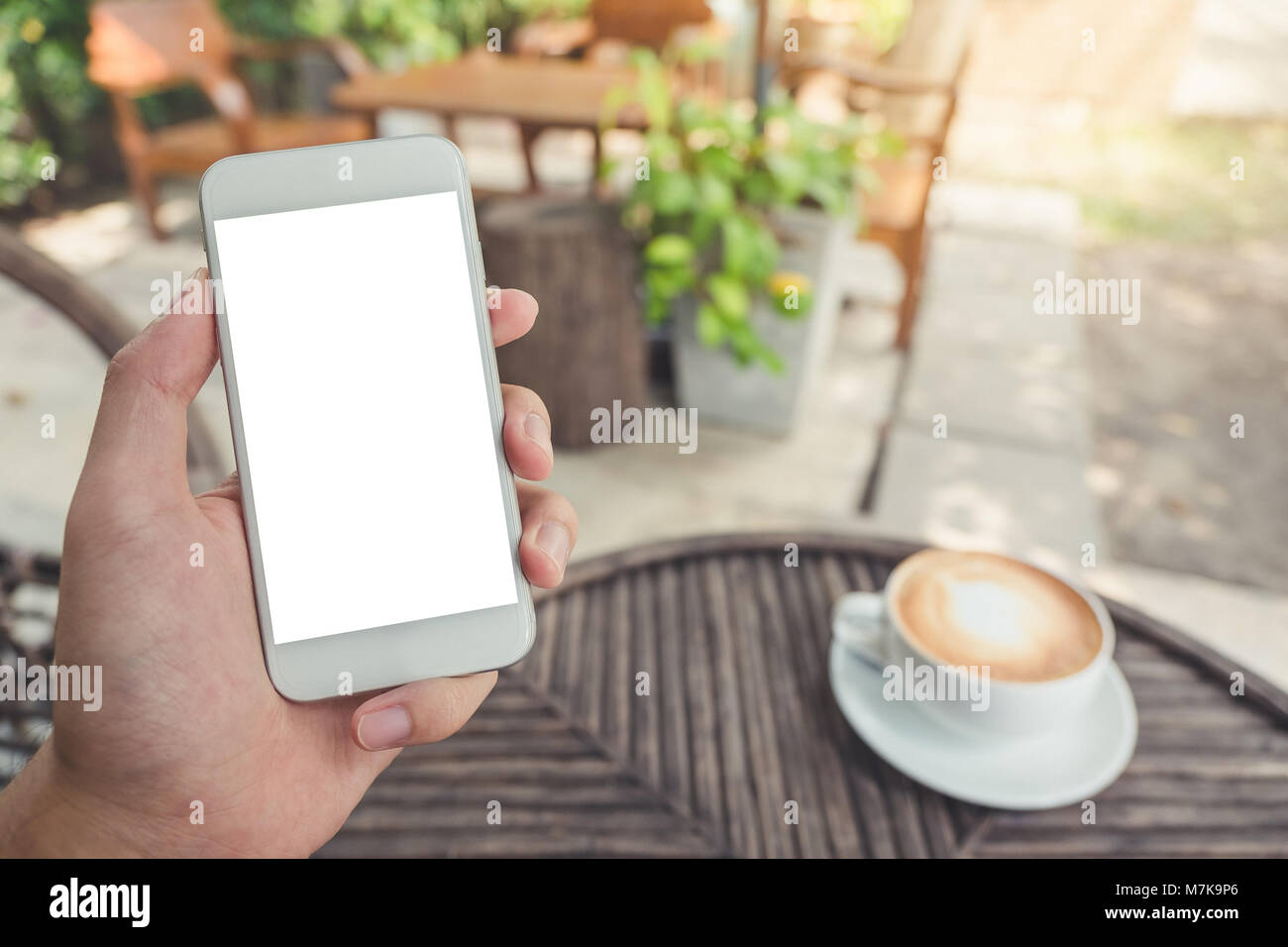 Immersive image de man's hand holding white téléphone mobile avec écran de bureau vierge dans un café avec l'arrière-plan flou outdoor Banque D'Images