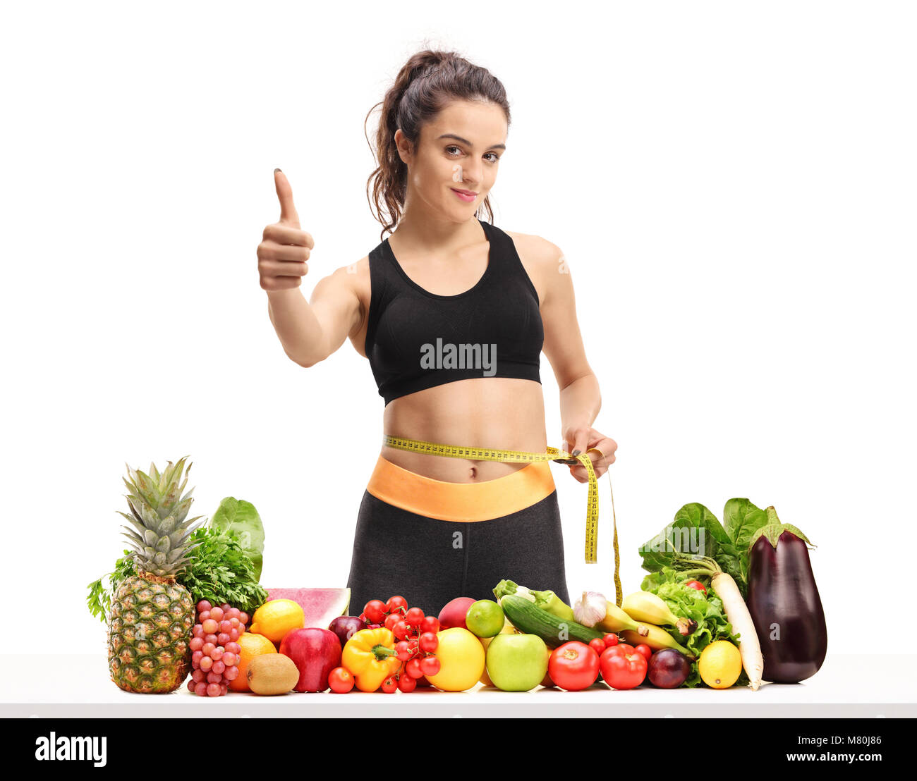 Girl measuring her waist remise en forme avec un ruban à mesurer et faire un pouce vers le haut signe derrière une table avec des fruits et légumes frais isolé sur blanc backgroun Banque D'Images