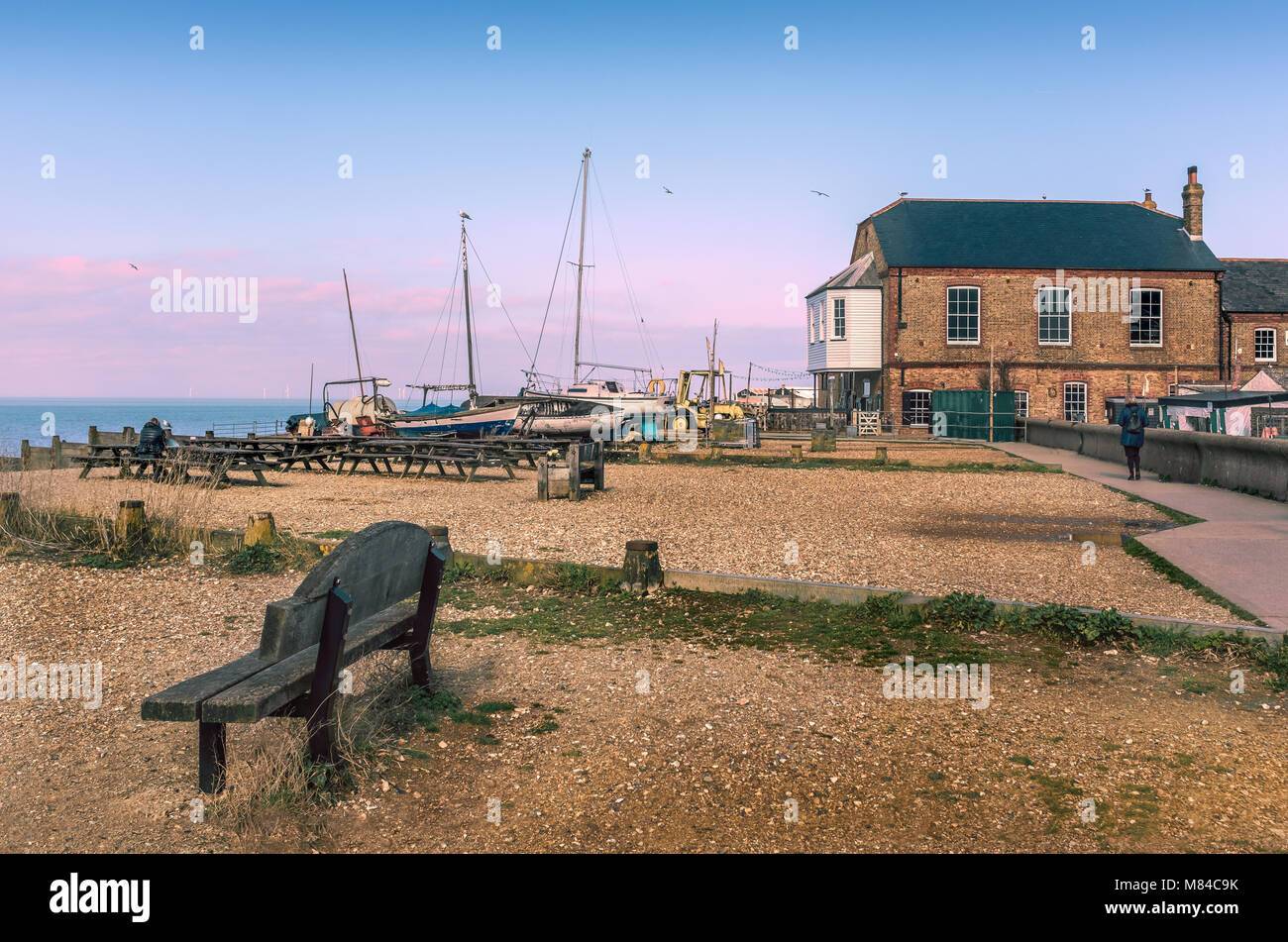Whitstable, Kent, UK Beach avec des bancs en bois et de vieux bateaux en bois amarré sur la plage. Il y a un bâtiment en brique rouge et un sentier pour piétons. Il y Banque D'Images