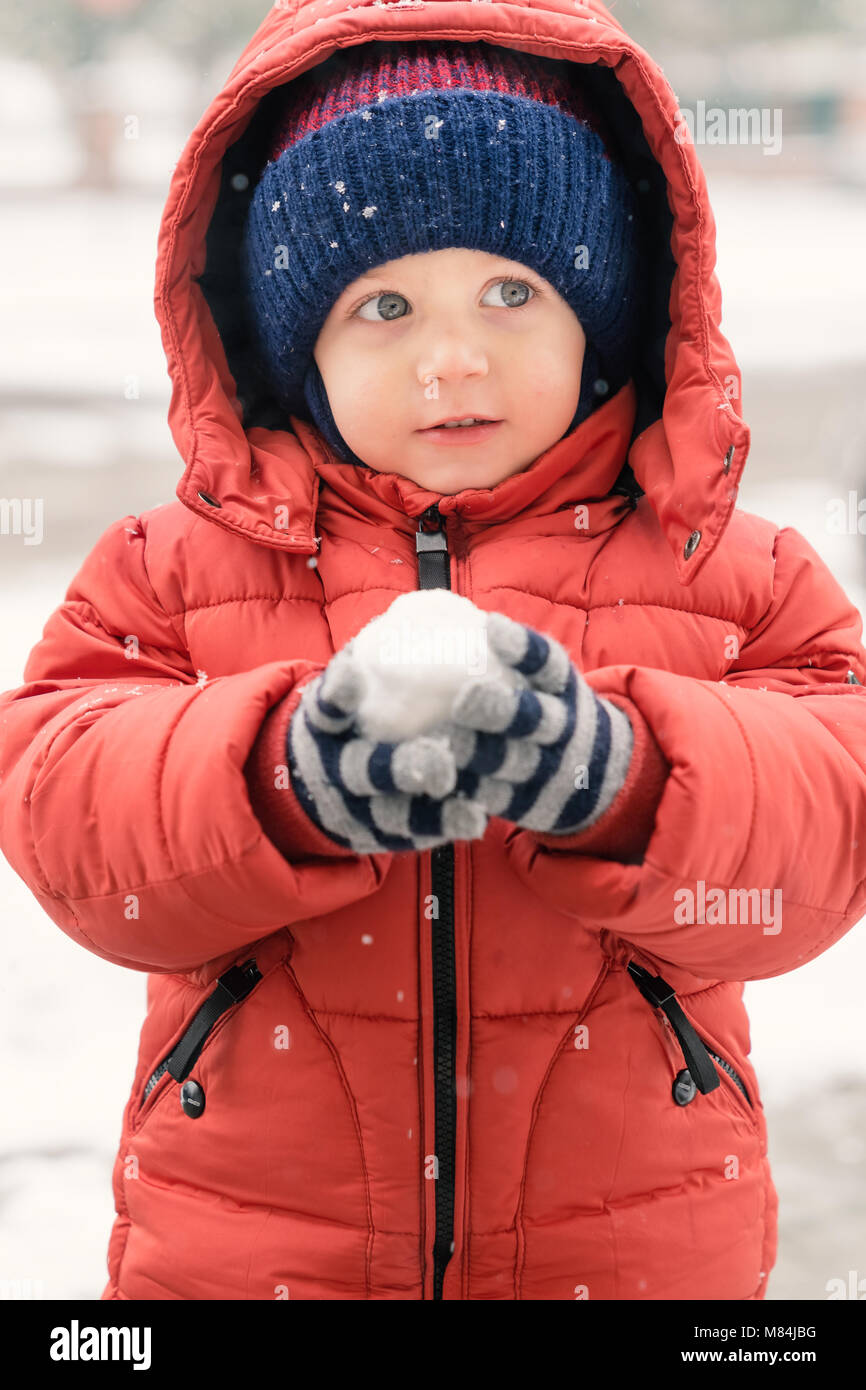 Beau Bébé garçon alors qu'il neige regarde vers le vide, tenant une boule  de neige dans ses mains, couvertes d'un manteau d'hiver rouge et un bonnet  de laine, c Photo Stock -
