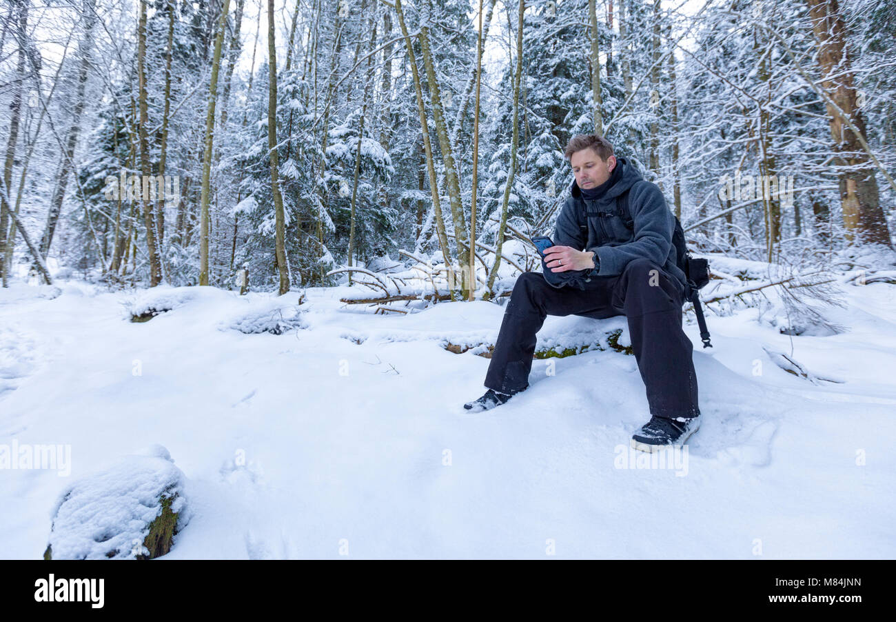 Homme adulte homme assis en bas dans la forêt en utilisant smartphone dans la neige paysage d'hiver modèle libération : Oui. Biens : Non. Banque D'Images