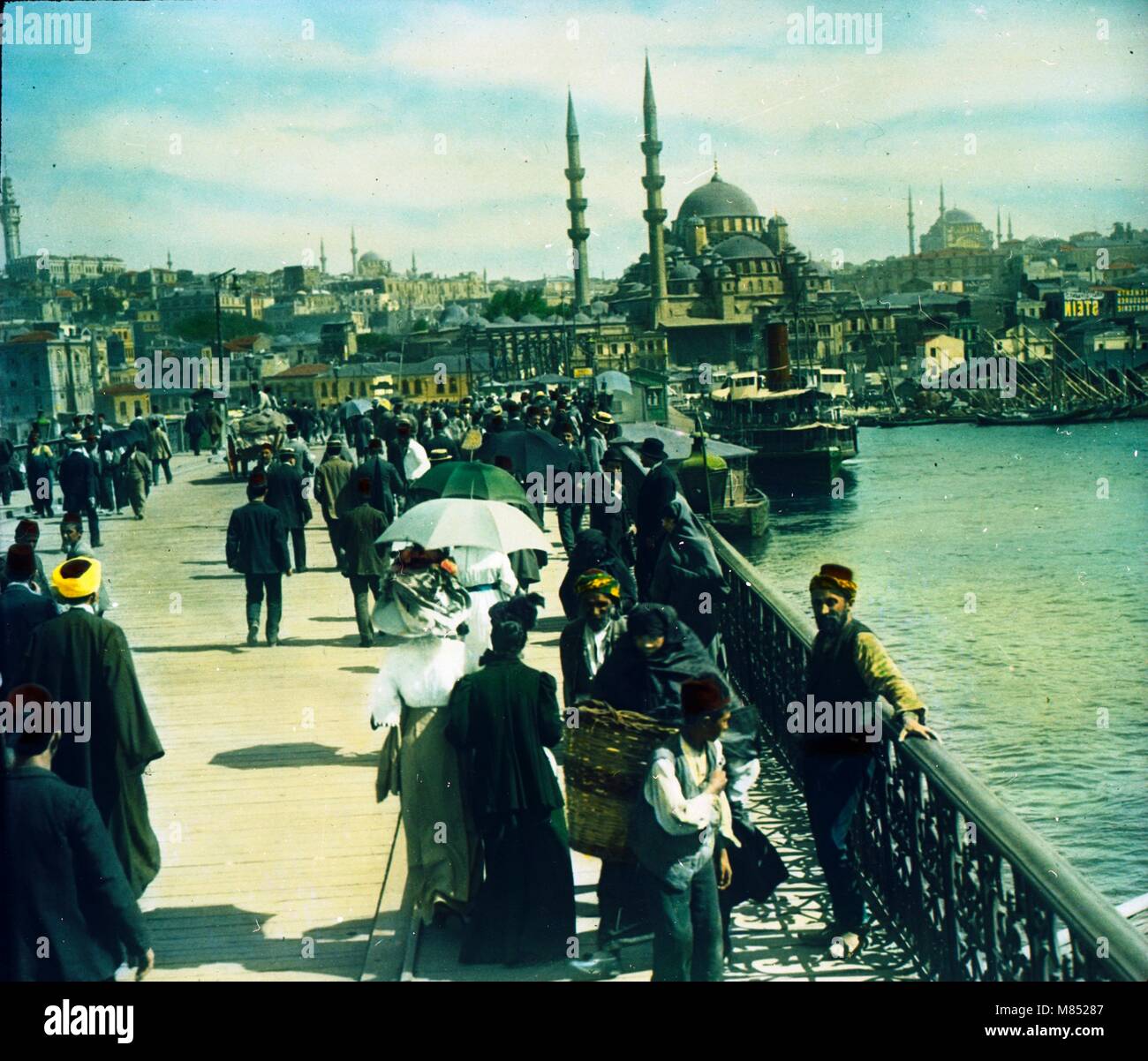 Vue colorée des piétons traversant le pont de Galata, avec la ligne d'horizon d'Istanbul en arrière-plan, Istanbul, Turquie, 1920. Au bout du pont de Galata se trouve la Nouvelle Mosquée. (Photo de Burton Holmes) Banque D'Images