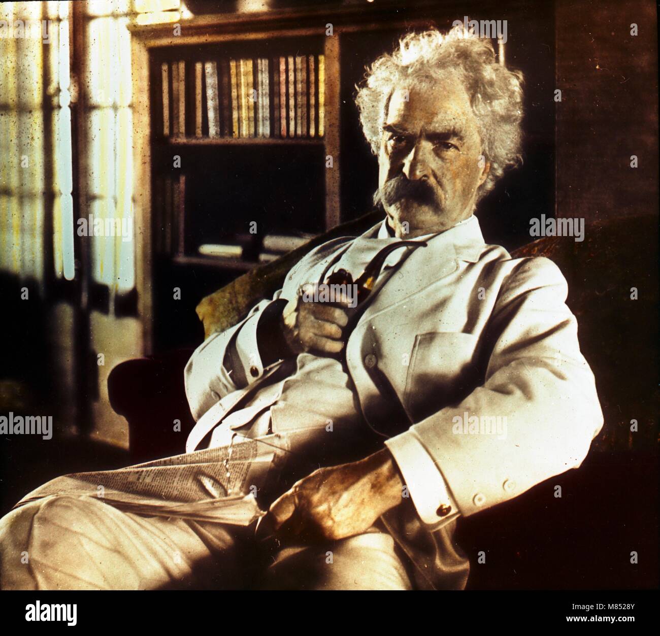 Portrait colorisé de l'auteur américain et humoriste Mark Twain, né Samuel Langhorne Clemens (1835 - 1910) tenant un tuyau et des verres, avec un journal sur ses genoux, vers 1900. (Photo de Burton Holmes) Banque D'Images