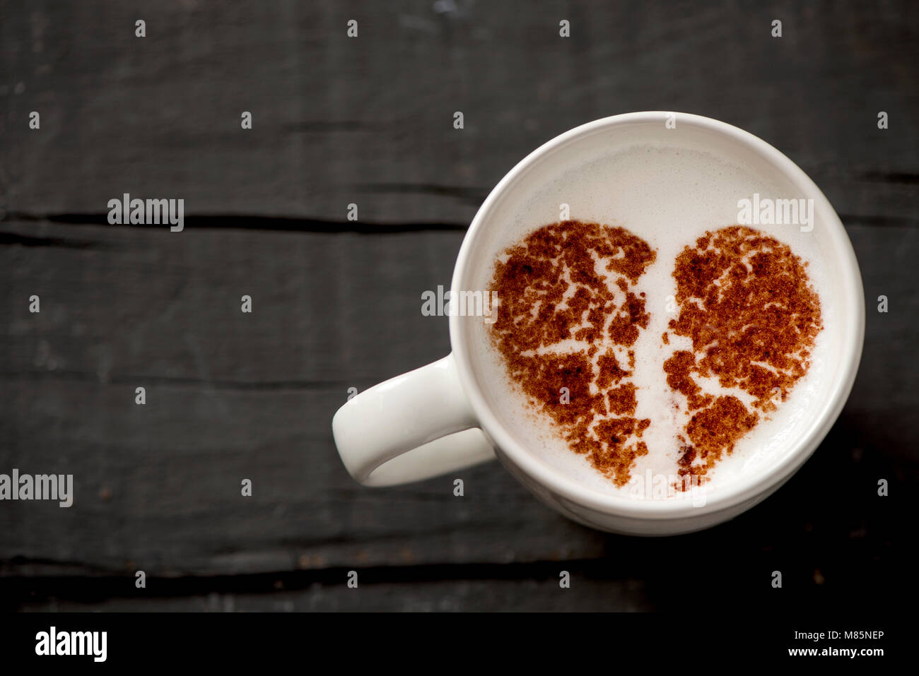 Capture d'un grand angle d'une tasse de Cappuccino en céramique blanche avec un cœur brisé dessiné avec du cacao en poudre sur sa mousse de lait, sur une table en bois rustique gris foncé Banque D'Images