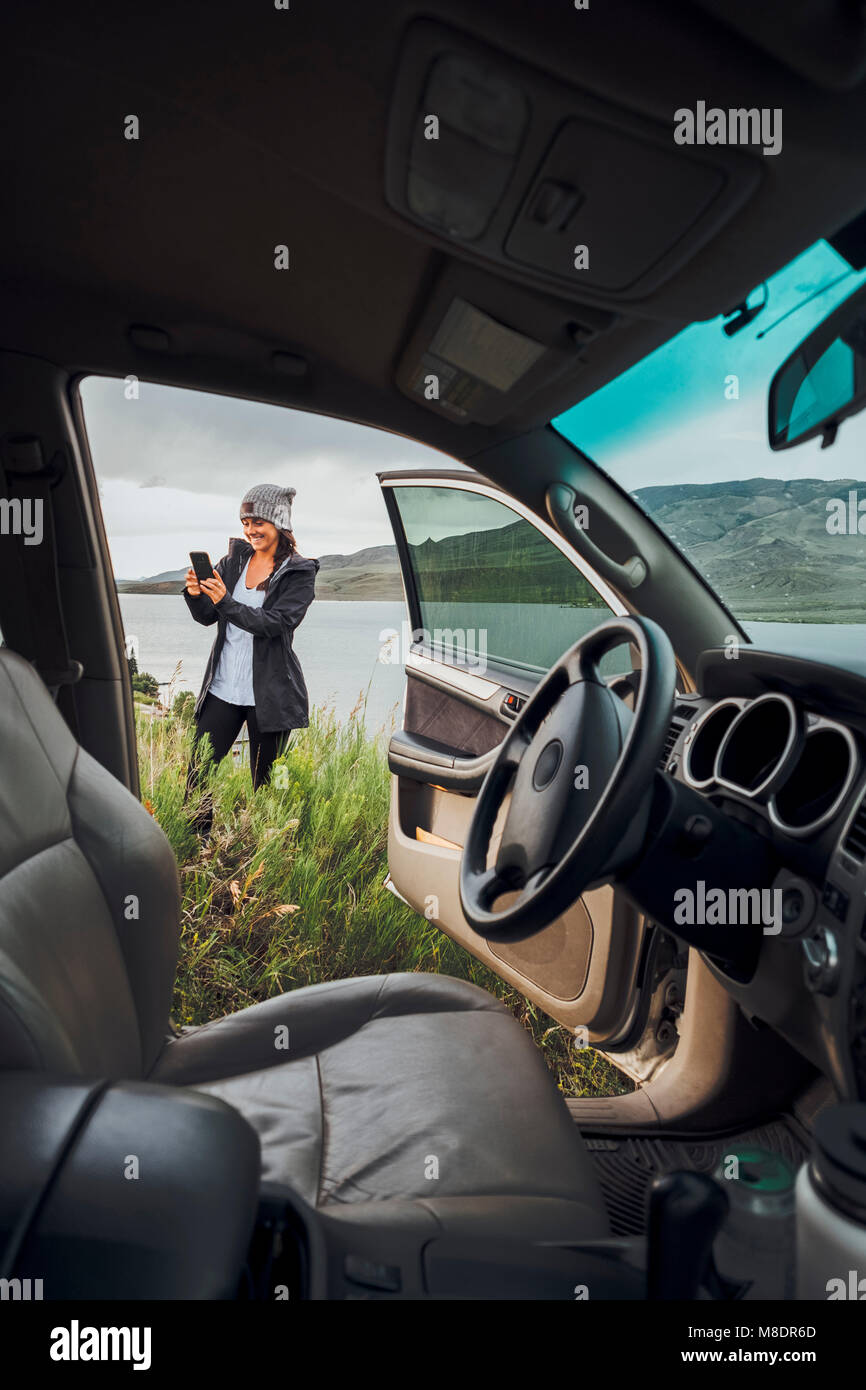 Jeune femme debout à côté du réservoir Dillon, holding smartphone, vue par voiture en stationnement, Silverthorne, Colorado, USA Banque D'Images