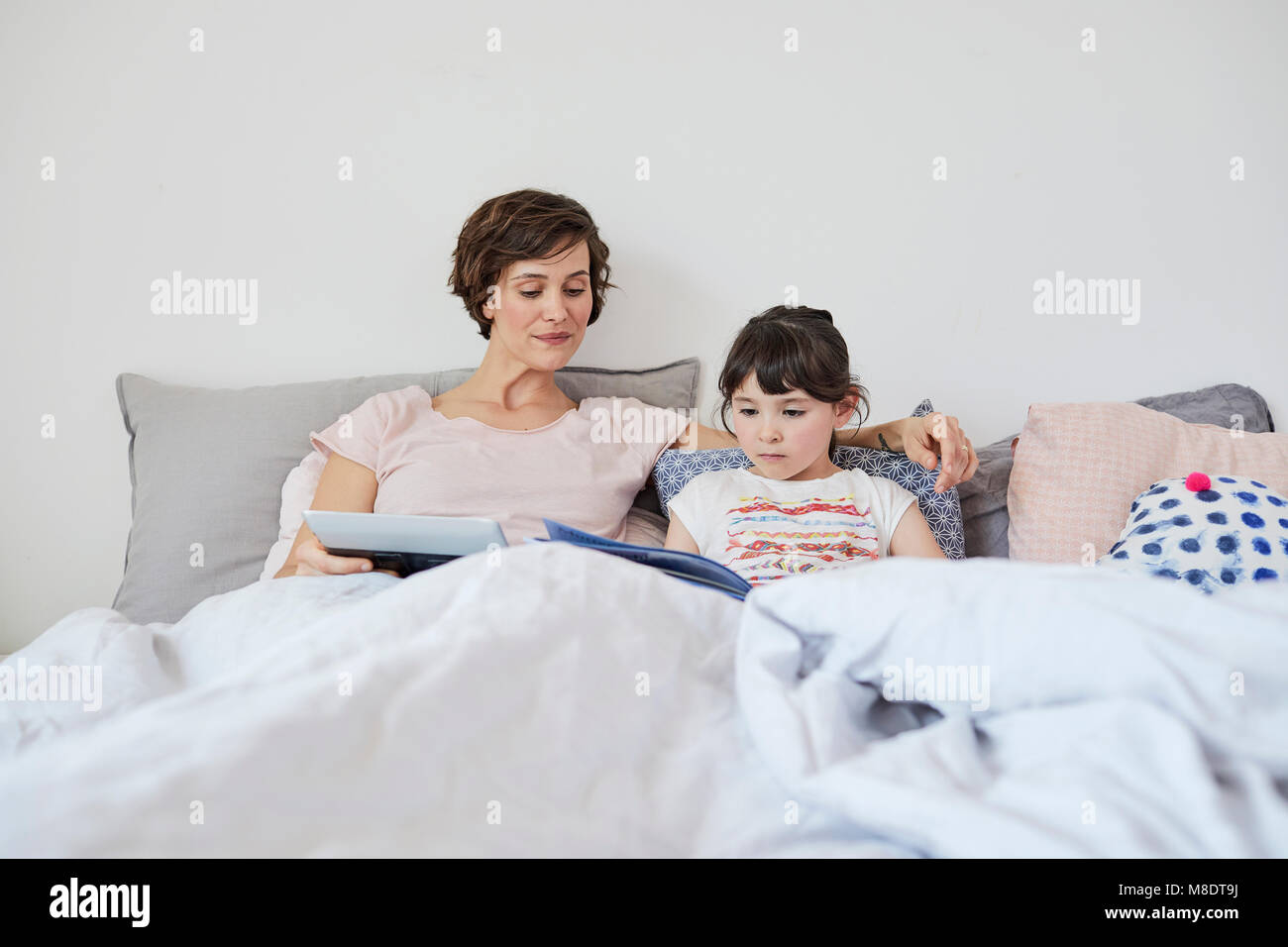 Mère et fille se détendre au lit, fille reading book, mère holding digital tablet Banque D'Images