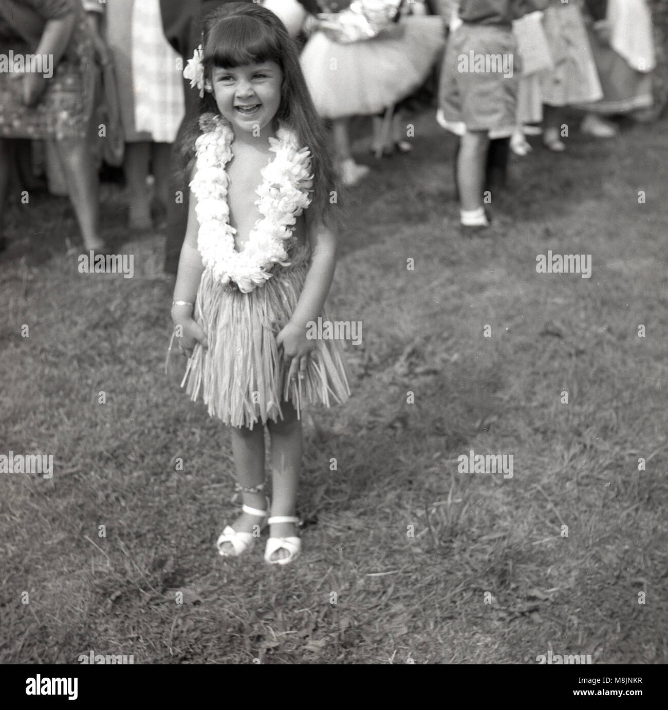 Années 1950, tableau historique, une petite fille heureuse fête de l'extérieur permanent smiling, portant fièrement son costume d'un hula ou jupe d'herbe avec collier de fleurs dans un bassin pour les concours costumé. Banque D'Images