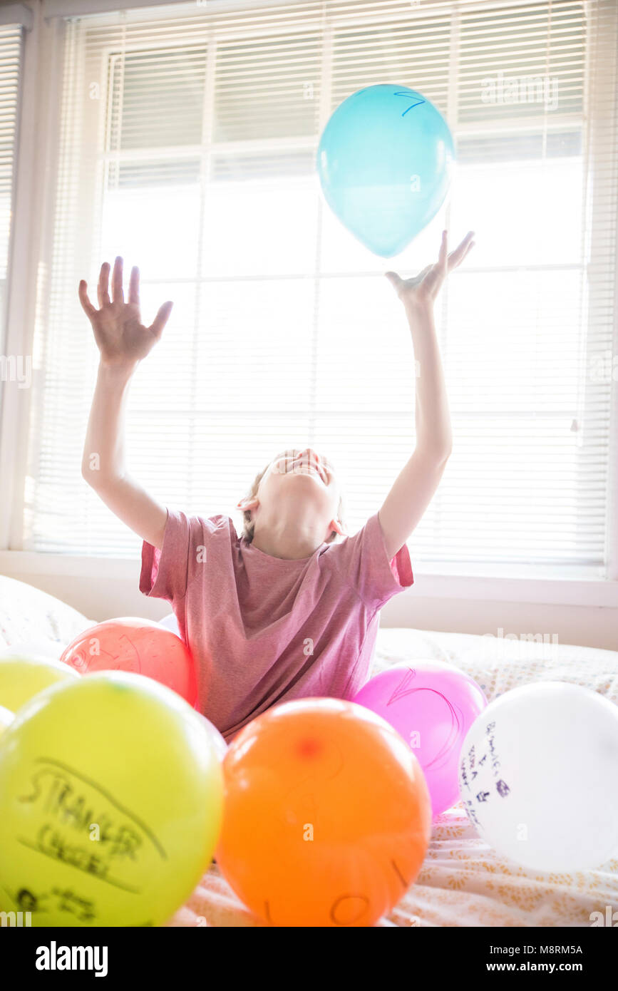 Happy boy playing with colorful balloons while sitting on bed at home Banque D'Images