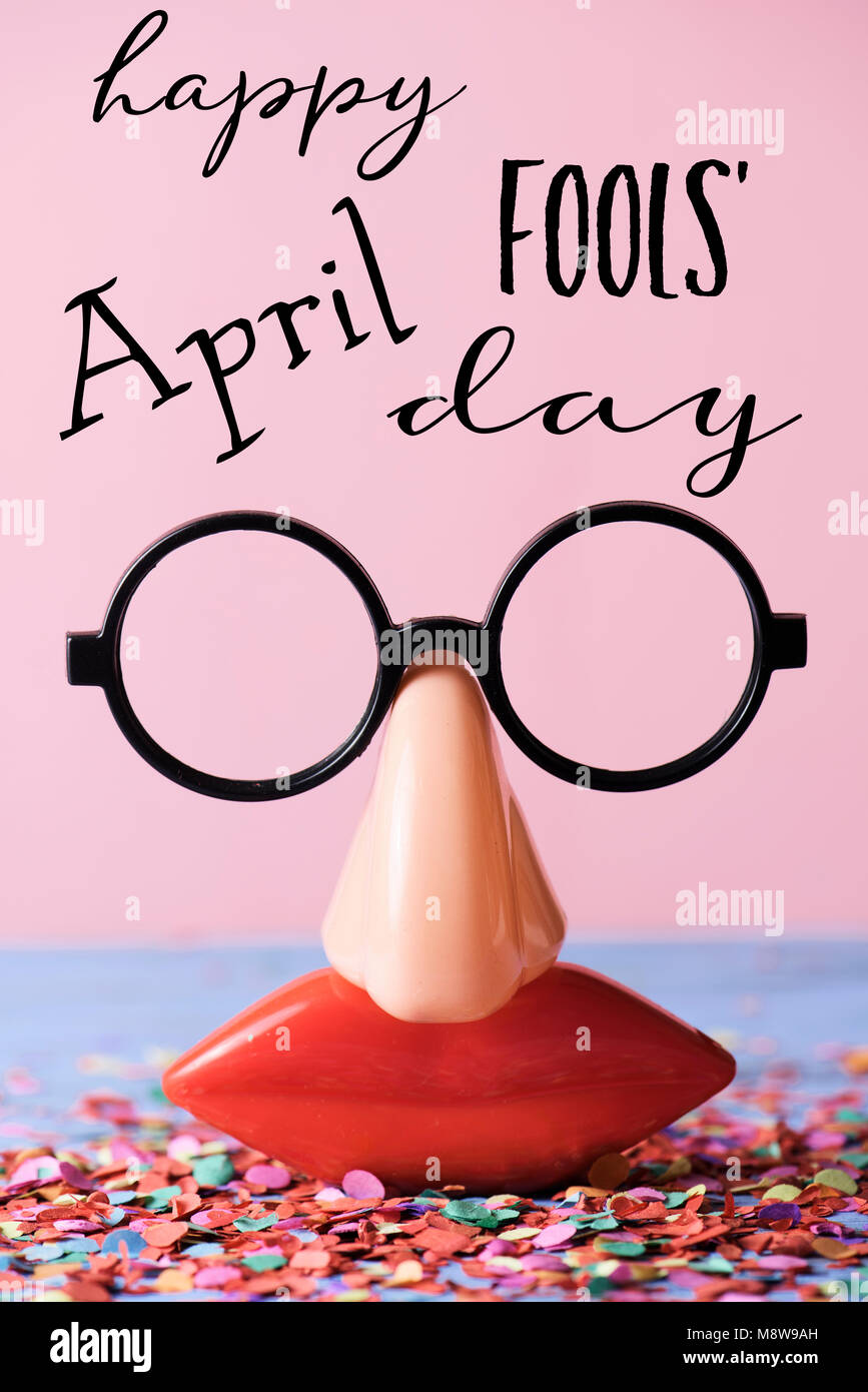 Une paire de lunettes, avec de faux nez et de la bouche, et l'happy April Fools Day, sur une surface rustique plein de confettis, contre un fond rose Banque D'Images