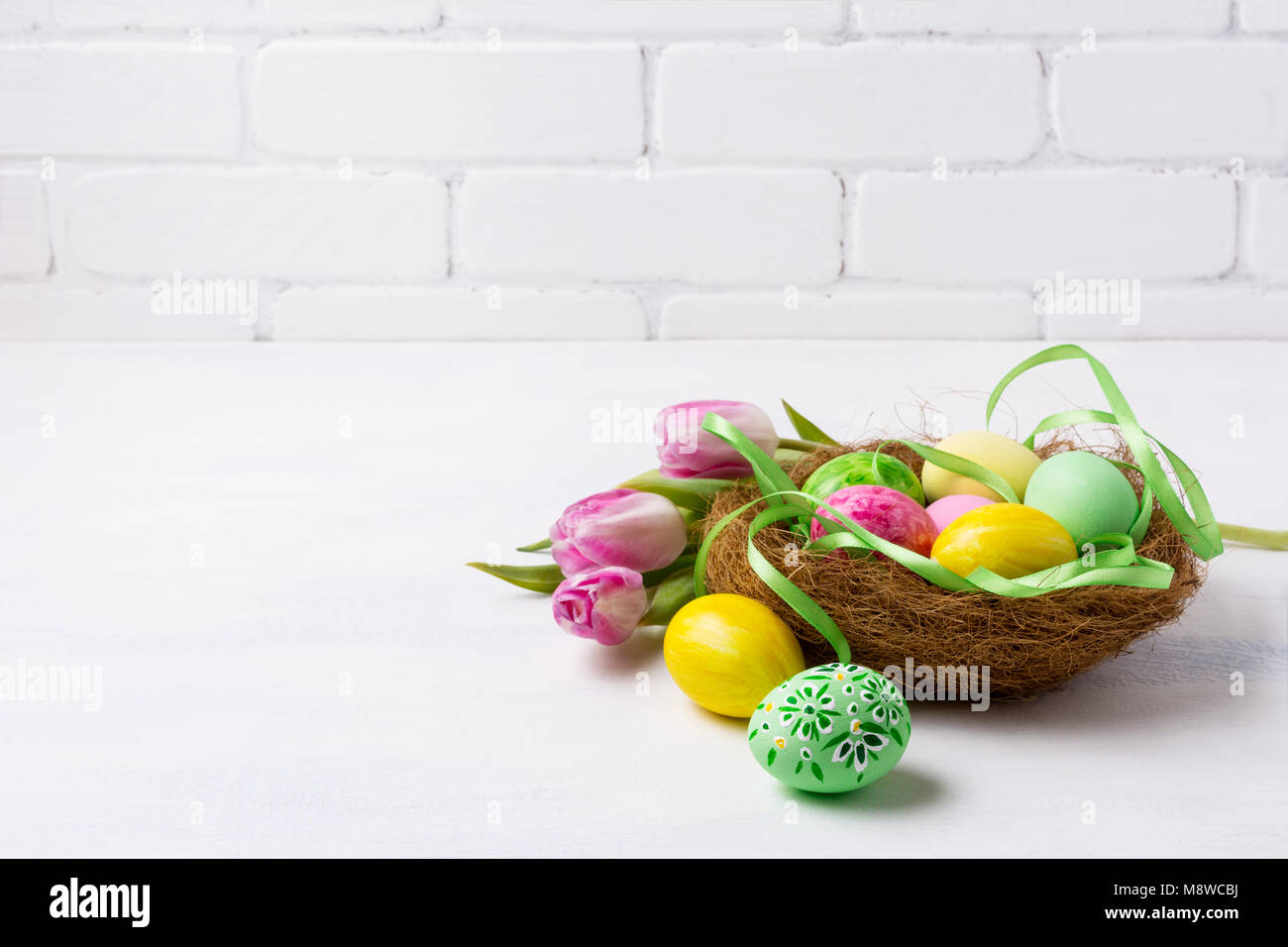 Joyeuses Pâques avec arrière-plan de message d'oeufs peints en vert et jaune dans le nid et des tulipes roses près de brick wall, copiez l'espace. Banque D'Images