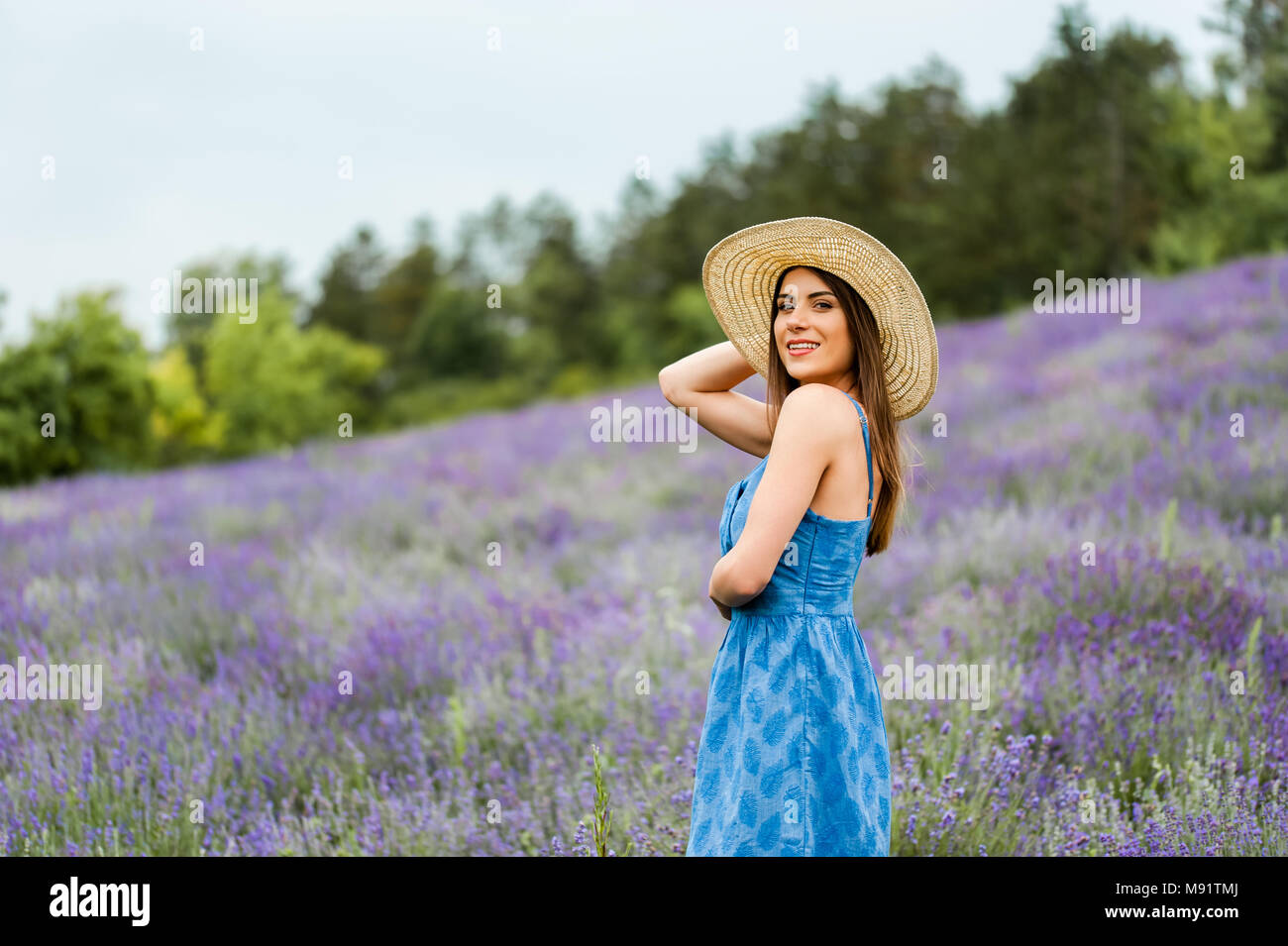 Femme au milieu d'un champ de lavande, vêtu d'une élégante robe bleue et un chapeau de paille, looking at camera posant avec un grand sourire et les mains. Banque D'Images