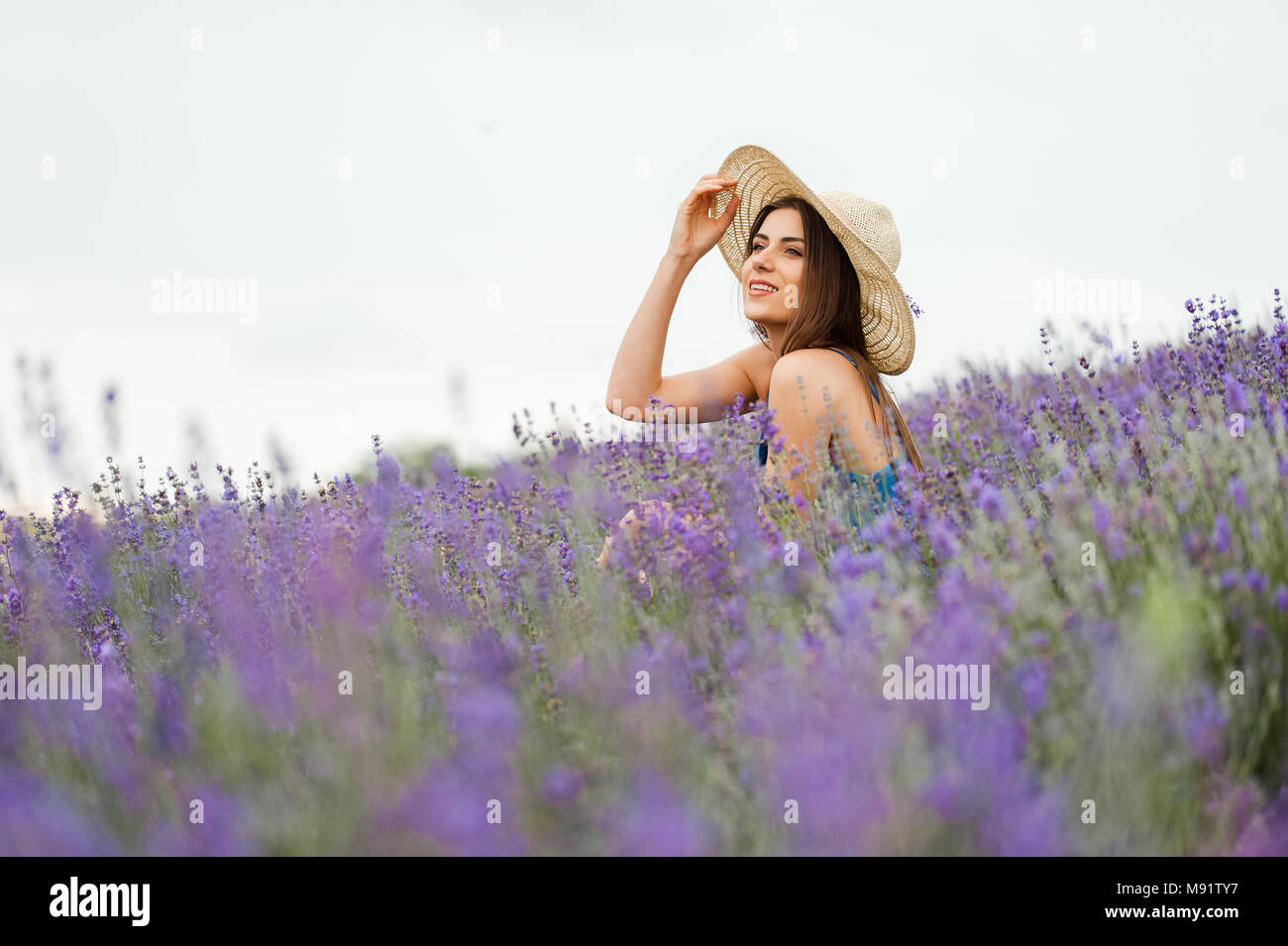 Femme au milieu d'un champ de lavande, vêtu d'une élégante robe bleue et un chapeau de paille, looking at camera posant avec un grand sourire et les mains. Banque D'Images