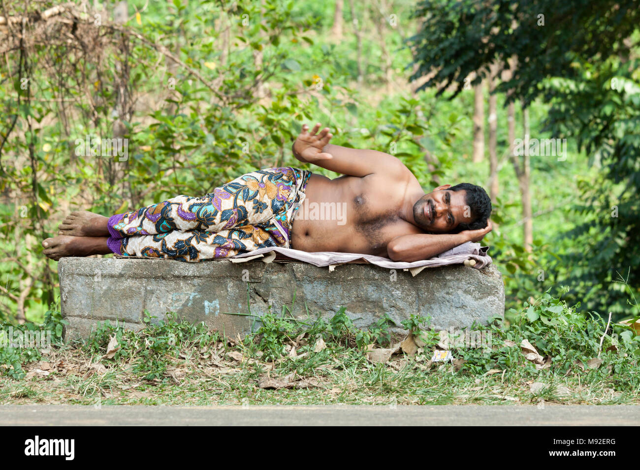 L'homme indien couché sur un bloc de béton vêtu d'un dhoti colorés et en agitant, Kerala, Inde. Banque D'Images